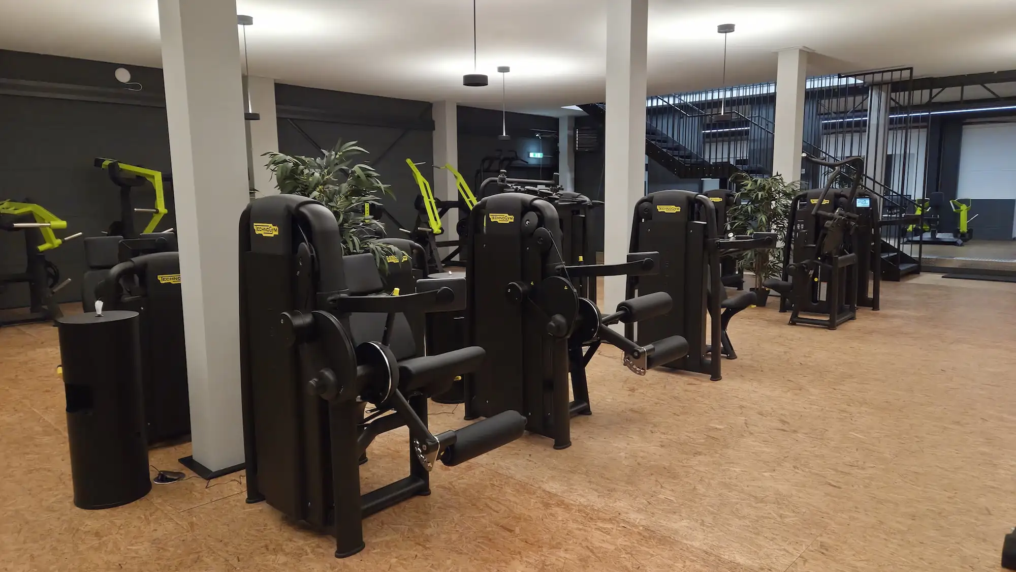 Moderner Fitnessraum mit verschiedenen schwarzen und gelben Technogym-Geräten auf hellem Holzboden und Wandbildern von trainierenden Personen.