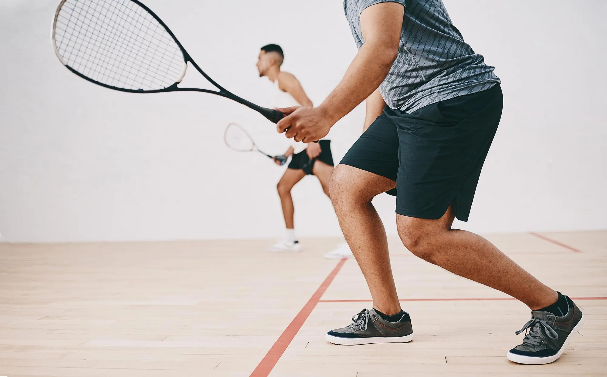 Zwei Männer spielen Squash in einem Hallen-Squashcourt, einer steht nah und hält den Schläger bereit.