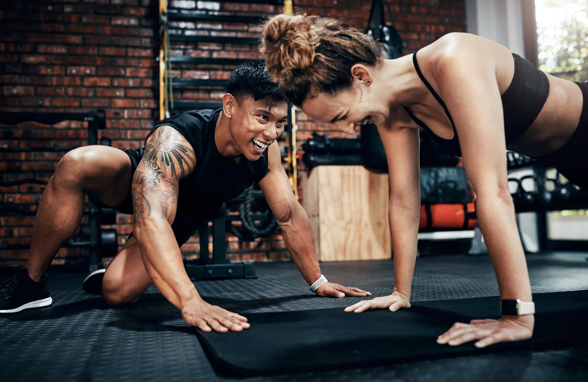 Zwei Personen, ein Mann und eine Frau, lachen und machen gemeinsam Liegestütze auf einer schwarzen Matte in einem Fitnessstudio.