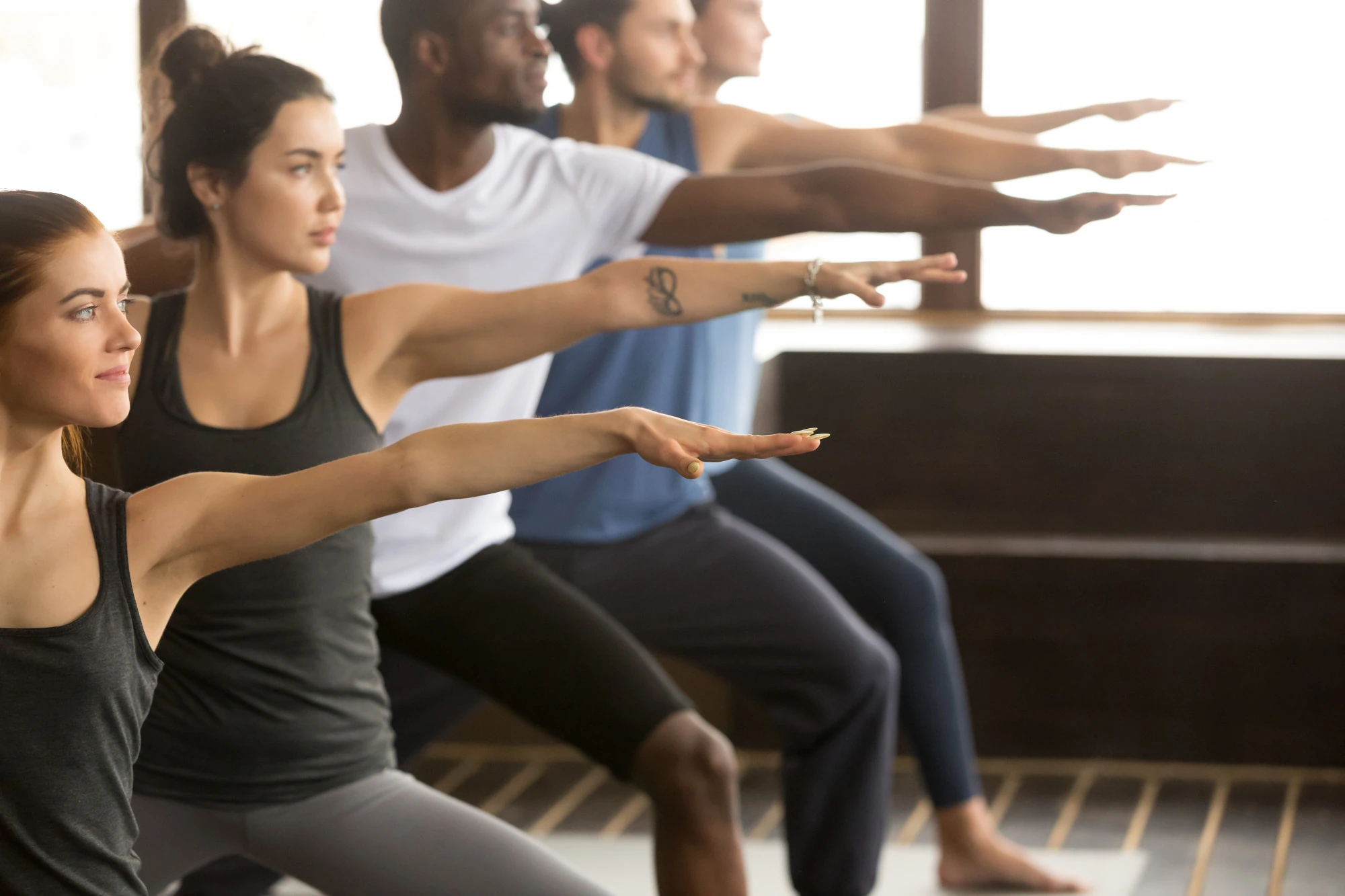 Gruppe von Menschen in sportlicher Kleidung macht Yoga-Übung Kriegerpose in einem hellen Raum.