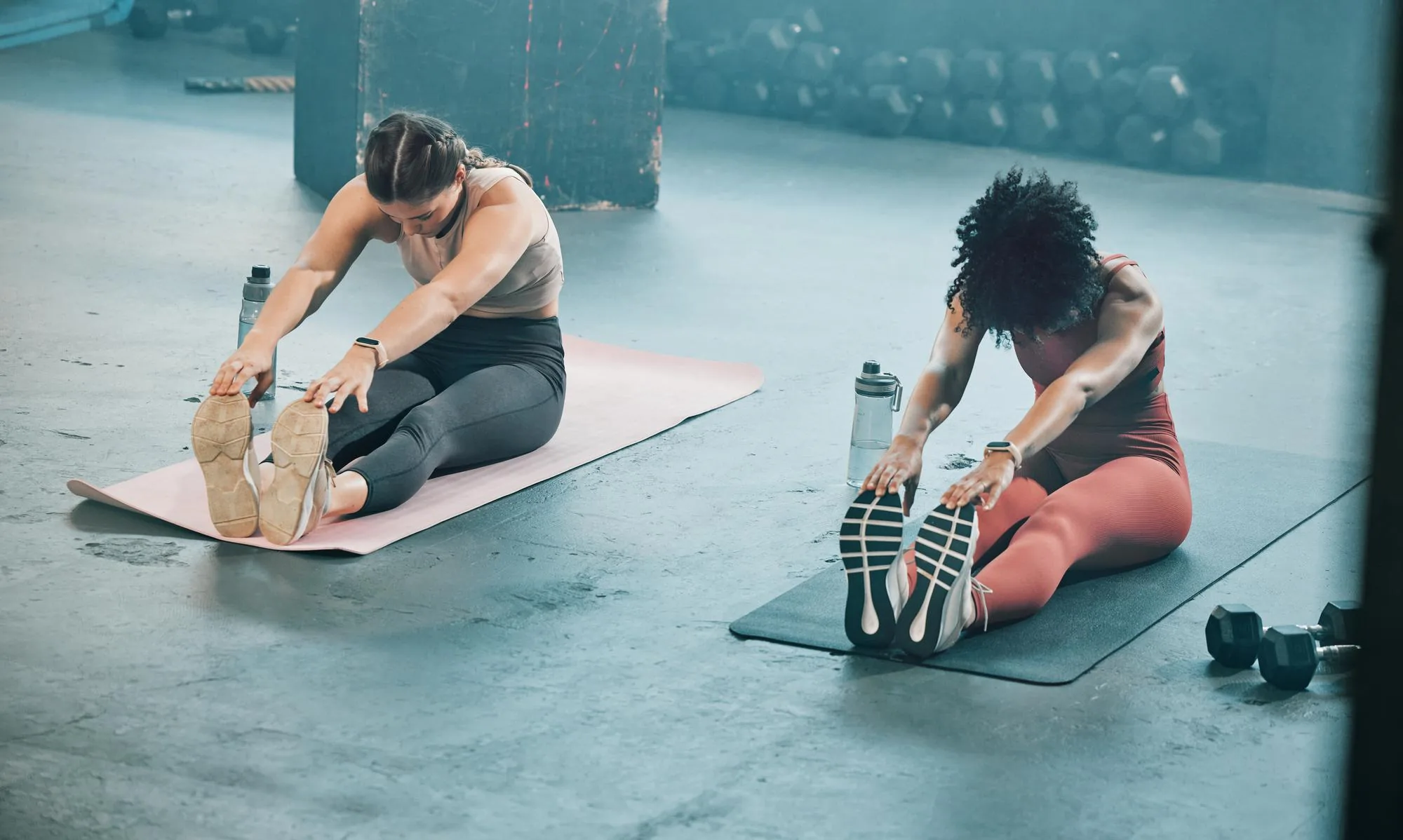 Zwei Frauen dehnen sich sitzend auf Yogamatten in einem Fitnessstudio, mit Wasserflaschen neben sich.