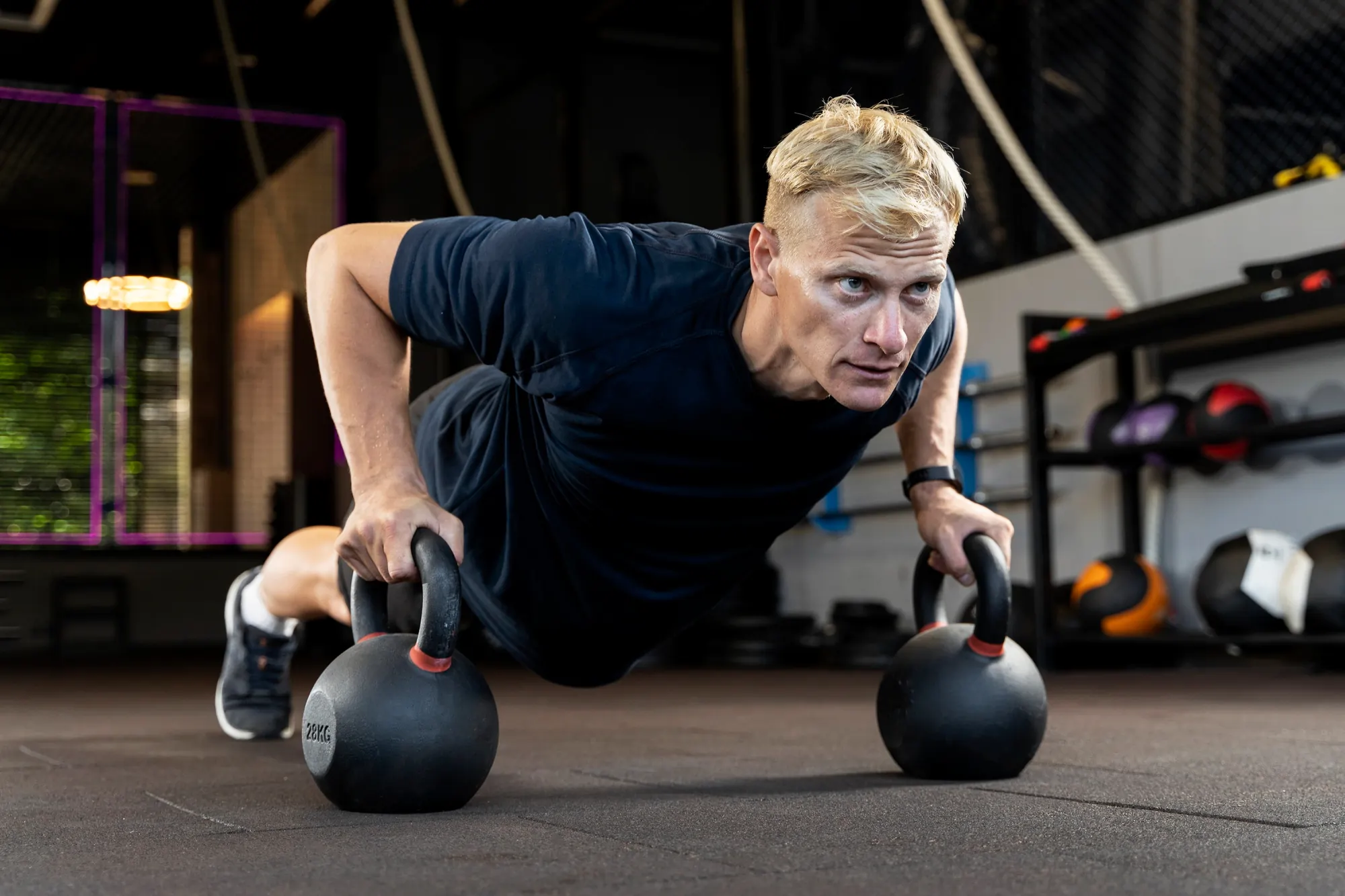 Mann macht Liegestütze mit beiden Händen auf schwarzen Kettlebells im Fitnessstudio.