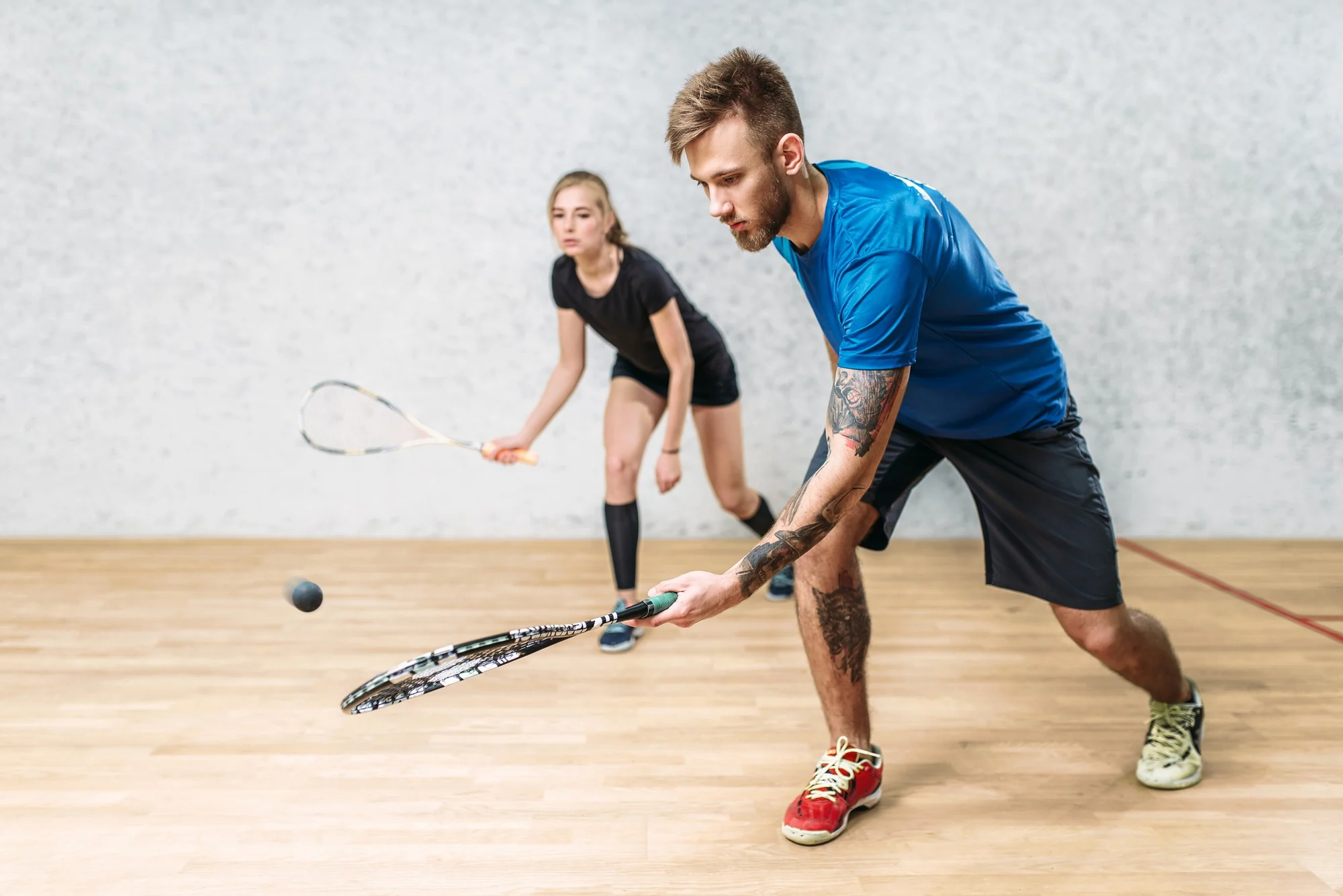 Zwei Personen spielen Squash in einem Hallenraum mit Holzboden, der Mann mit blauer Kleidung schlägt den Ball mit dem Schläger.