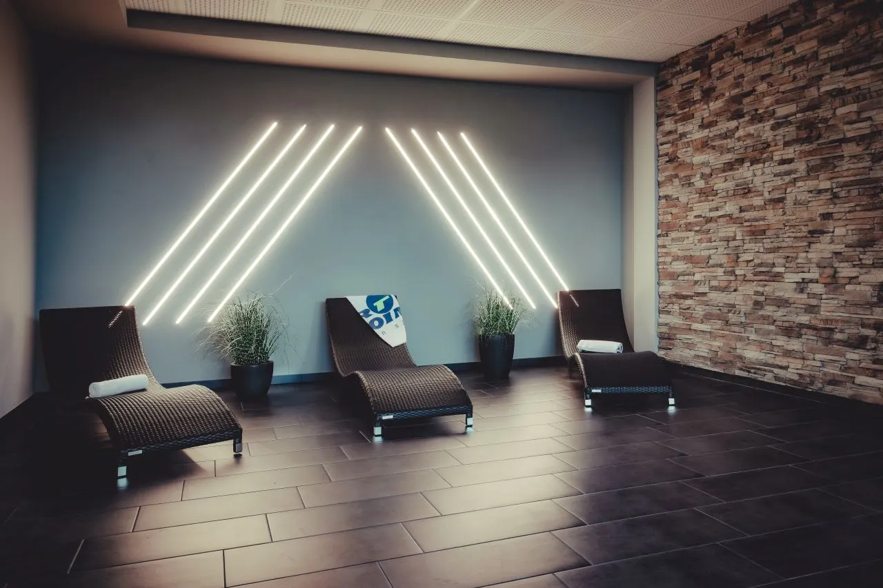 Drei braune Liegestühle mit weißen Handtüchern und Pflanzen an der Wand mit beleuchteten diagonalen Lichtstreifen in einem modernen Entspannungsraum.