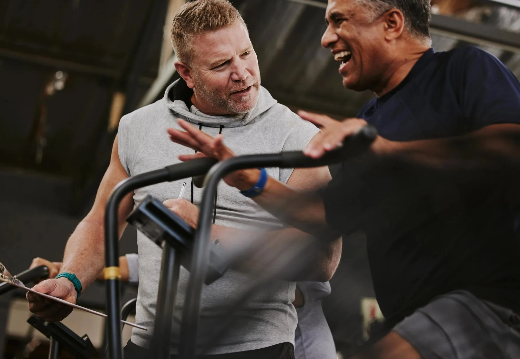 Zwei Männer im Fitnessstudio sprechen, einer trägt einen grauen Kapuzenpullover und hält ein Klemmbrett, der andere trainiert auf einem Fitnessgerät und lacht.