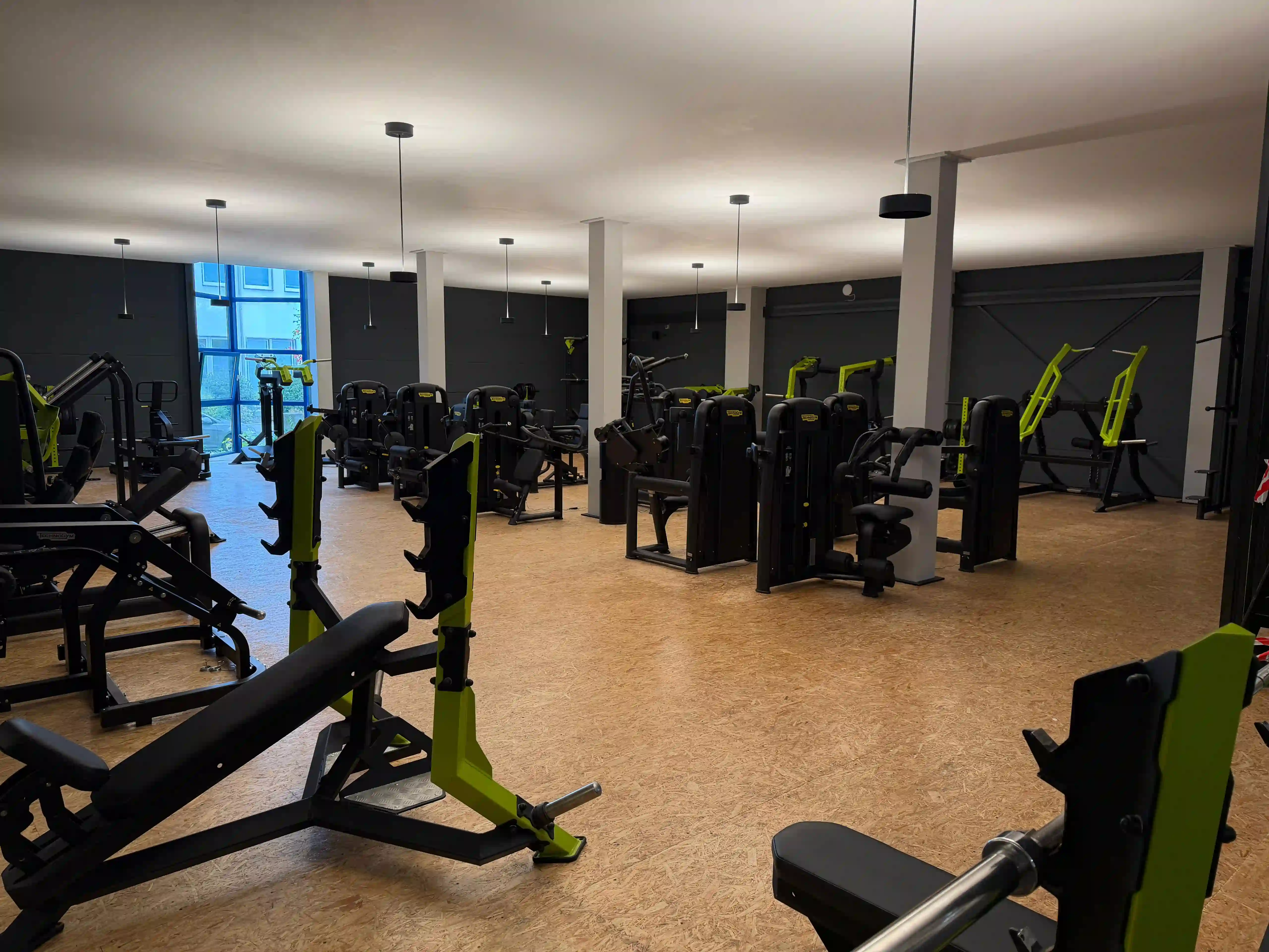 Großer Fitnessraum mit Krafttrainingsgeräten und Holzfußboden, beleuchtet von Deckenlampen und Tageslicht durch Fenster.