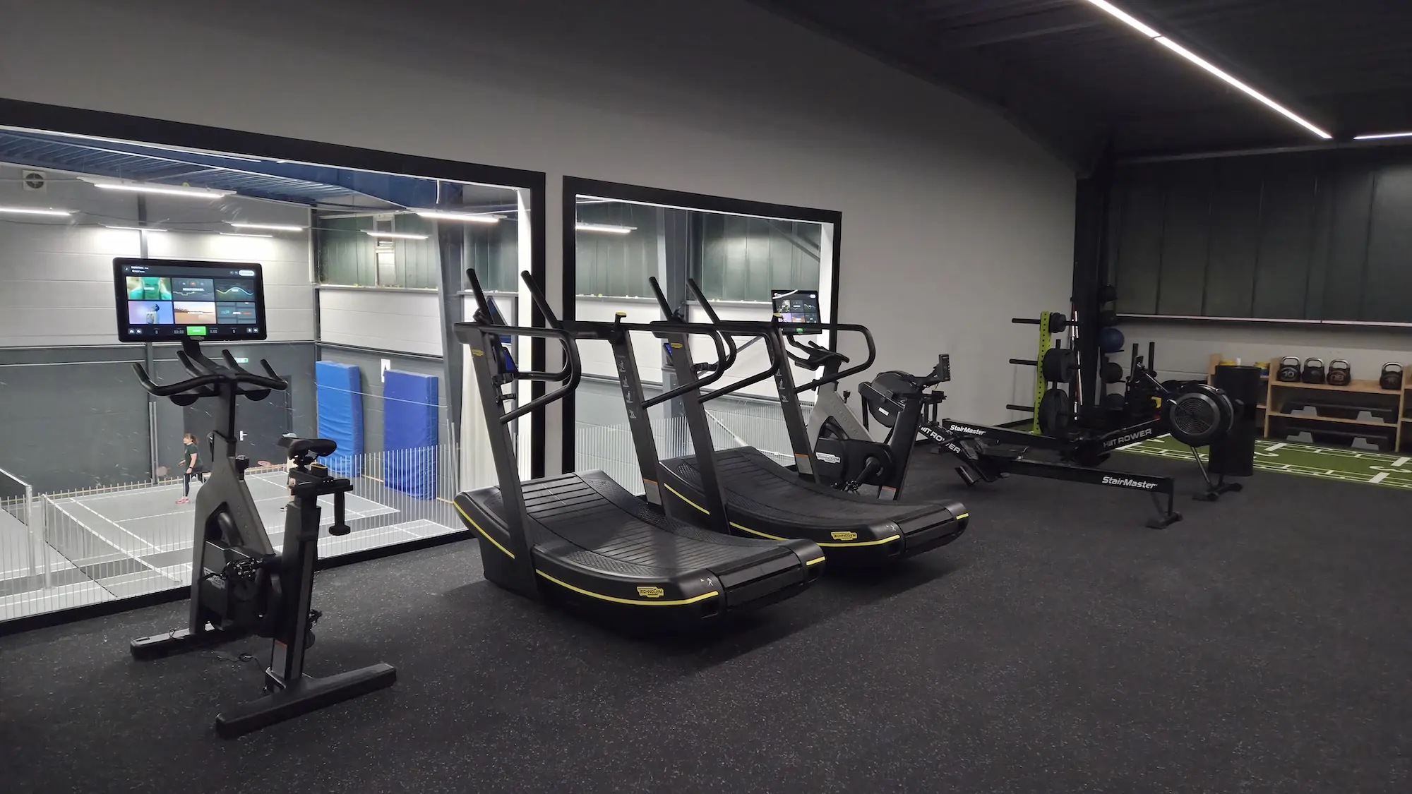 Moderne Fitnessgeräte in einem gut beleuchteten Fitnessstudio mit Blick auf eine Halle mit Sportler.