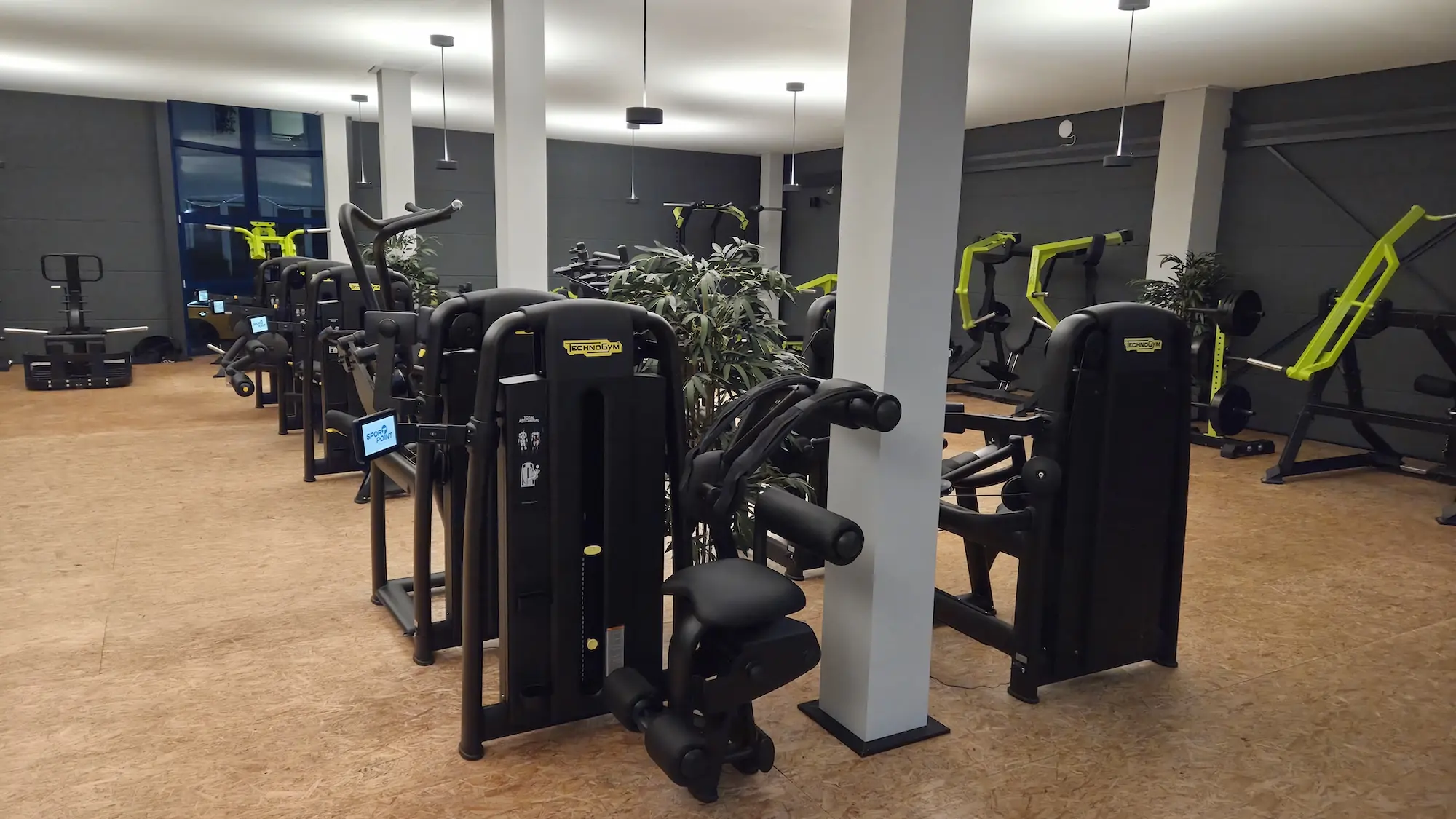Innenansicht eines Fitnessstudios mit verschiedenen schwarzen und grünen Trainingsgeräten auf einem hellen Holzfußboden.