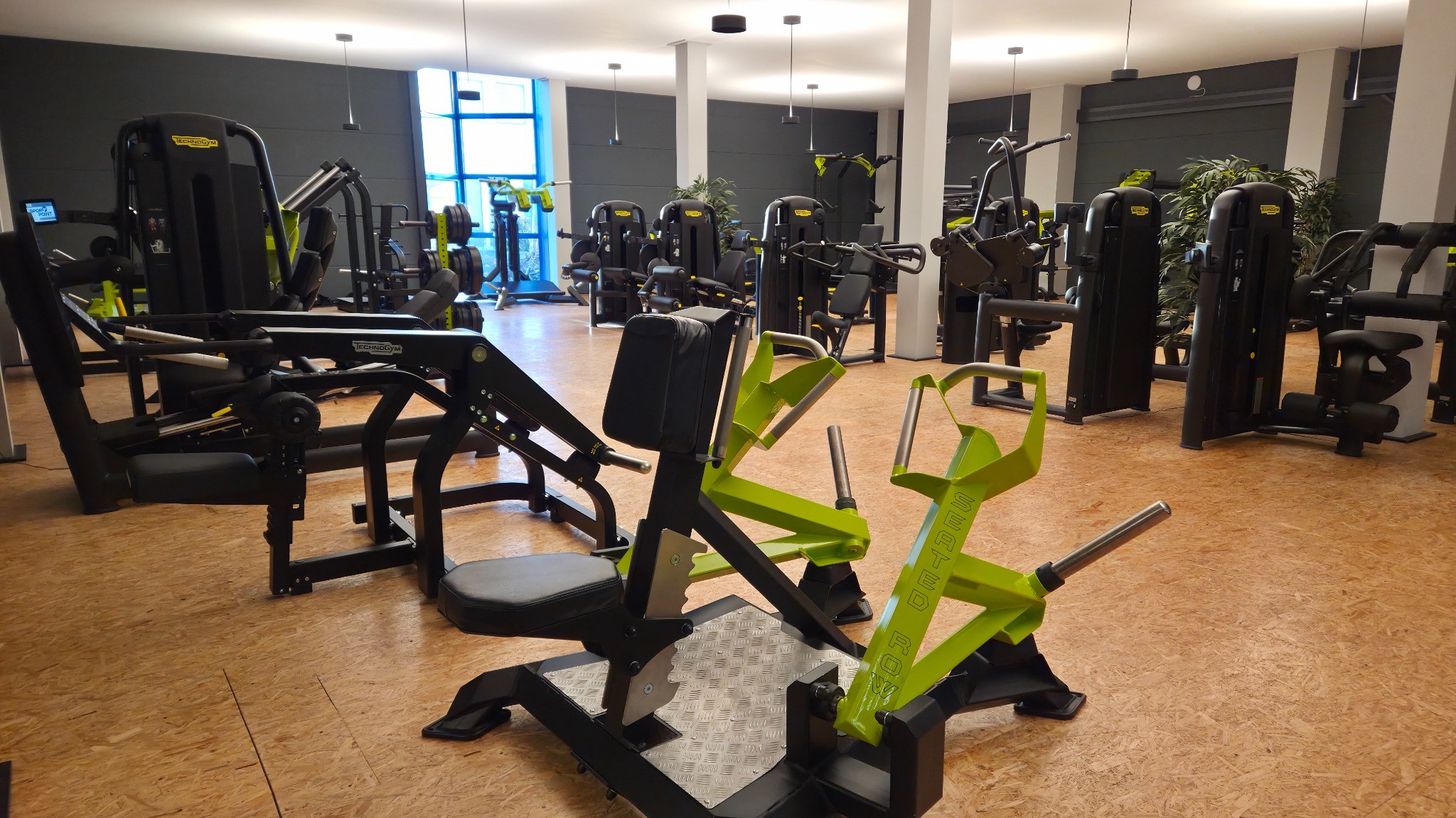 Leerer Fitnessraum mit verschiedenen Trainingsgeräten auf Holzboden und großen Fenstern.