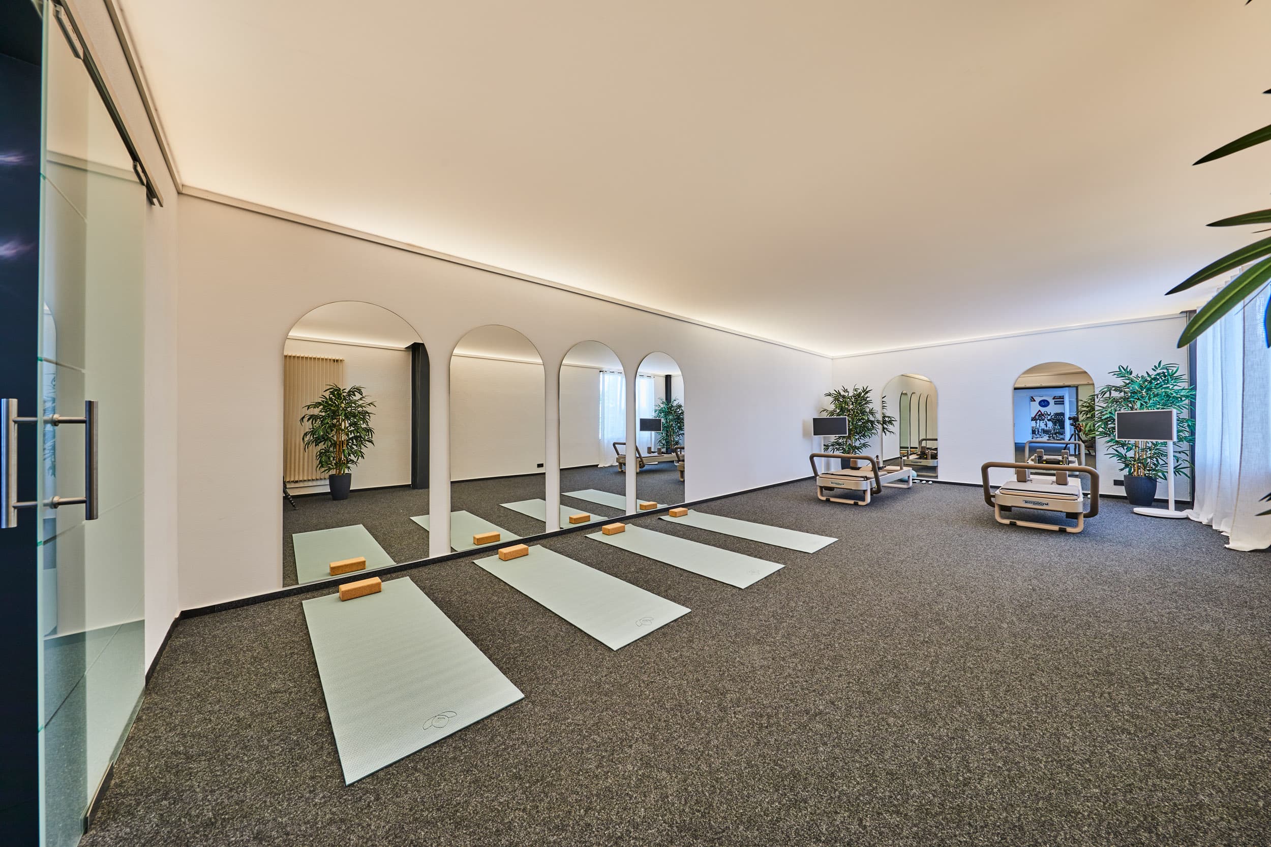 Heller, moderner Übungsraum mit drei blauen Yogamatten und Blöcken auf dem Teppichboden vor einer Wand mit vier großen, bogenförmigen Spiegeln, zwei Pilates-Geräten mit Monitoren, mehreren großen Pflanzen und einem hellen, indirekten Deckenlicht.
