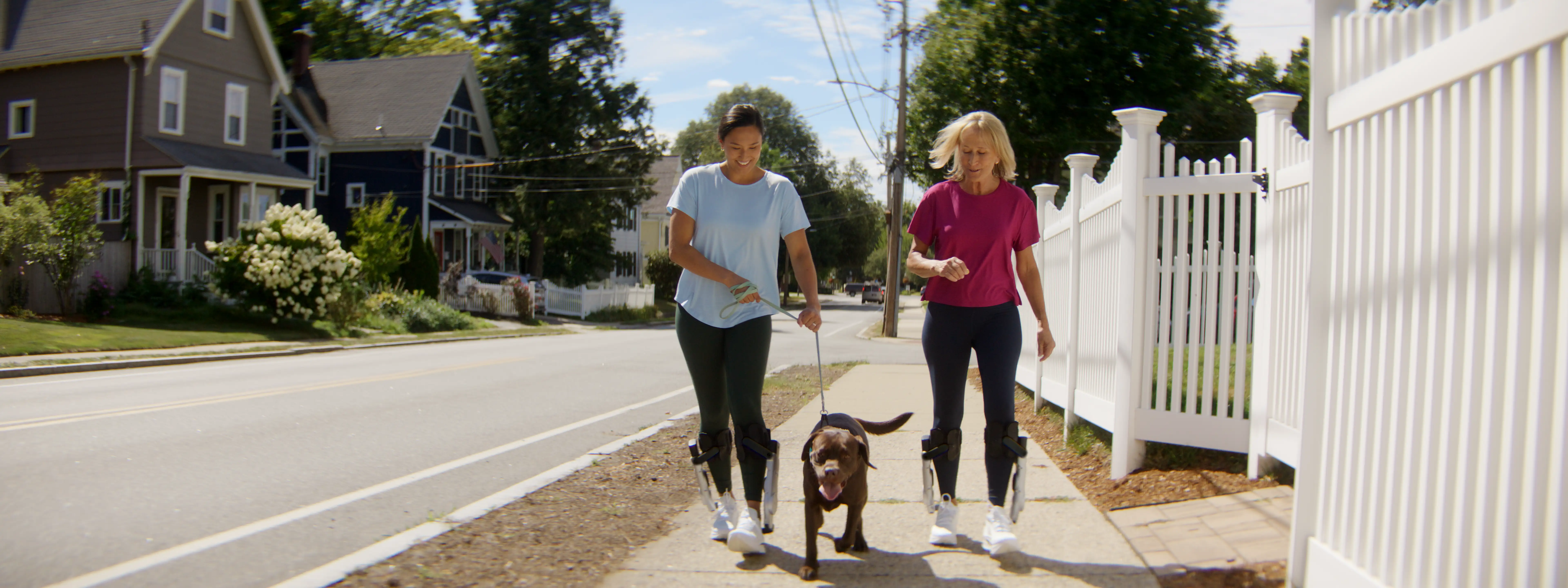 Two women walking together on a suburban sidewalk with a brown dog on a leash