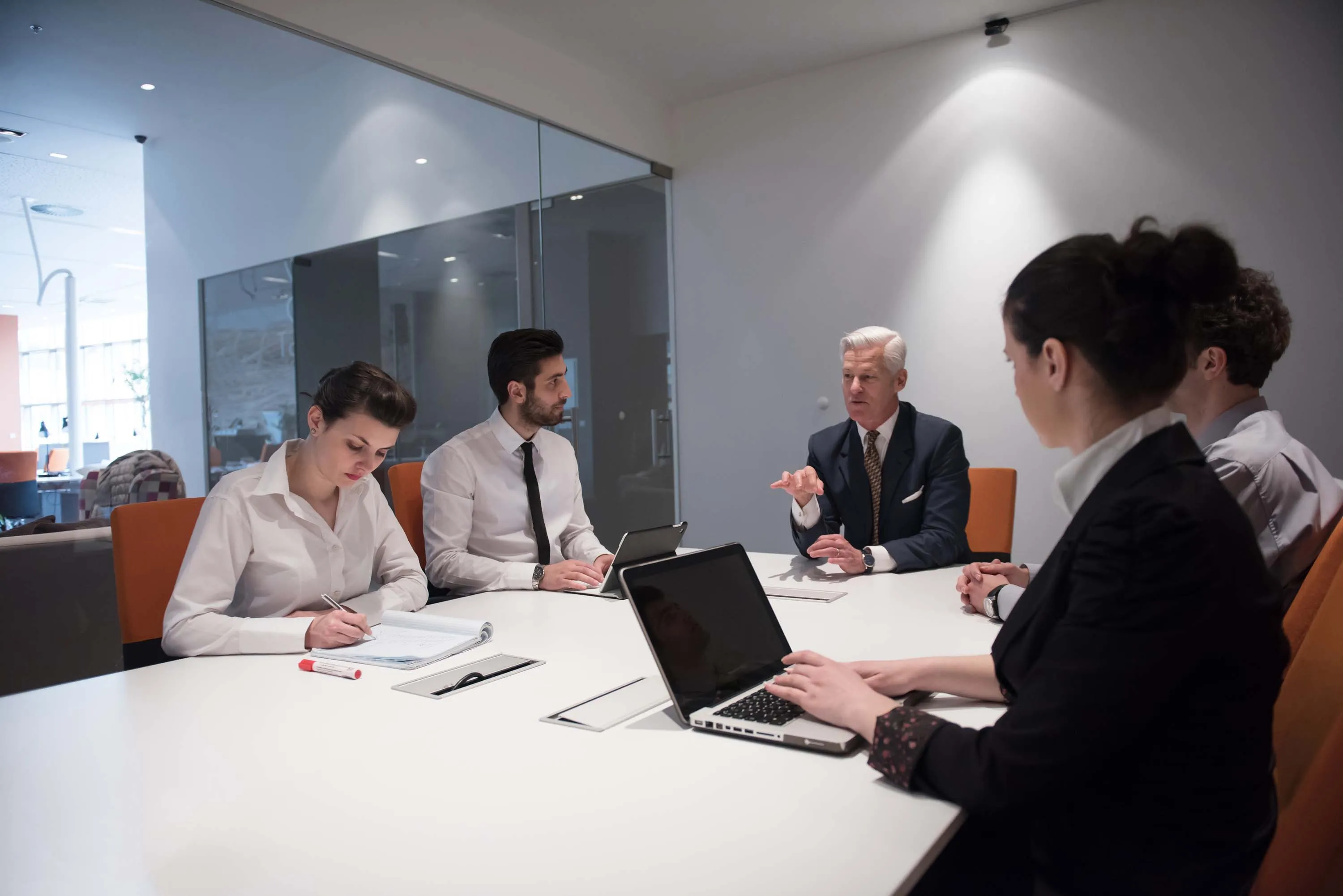 Business meeting with five professionals seated around a conference table, one older man in a suit speaking while others take notes and use laptops.