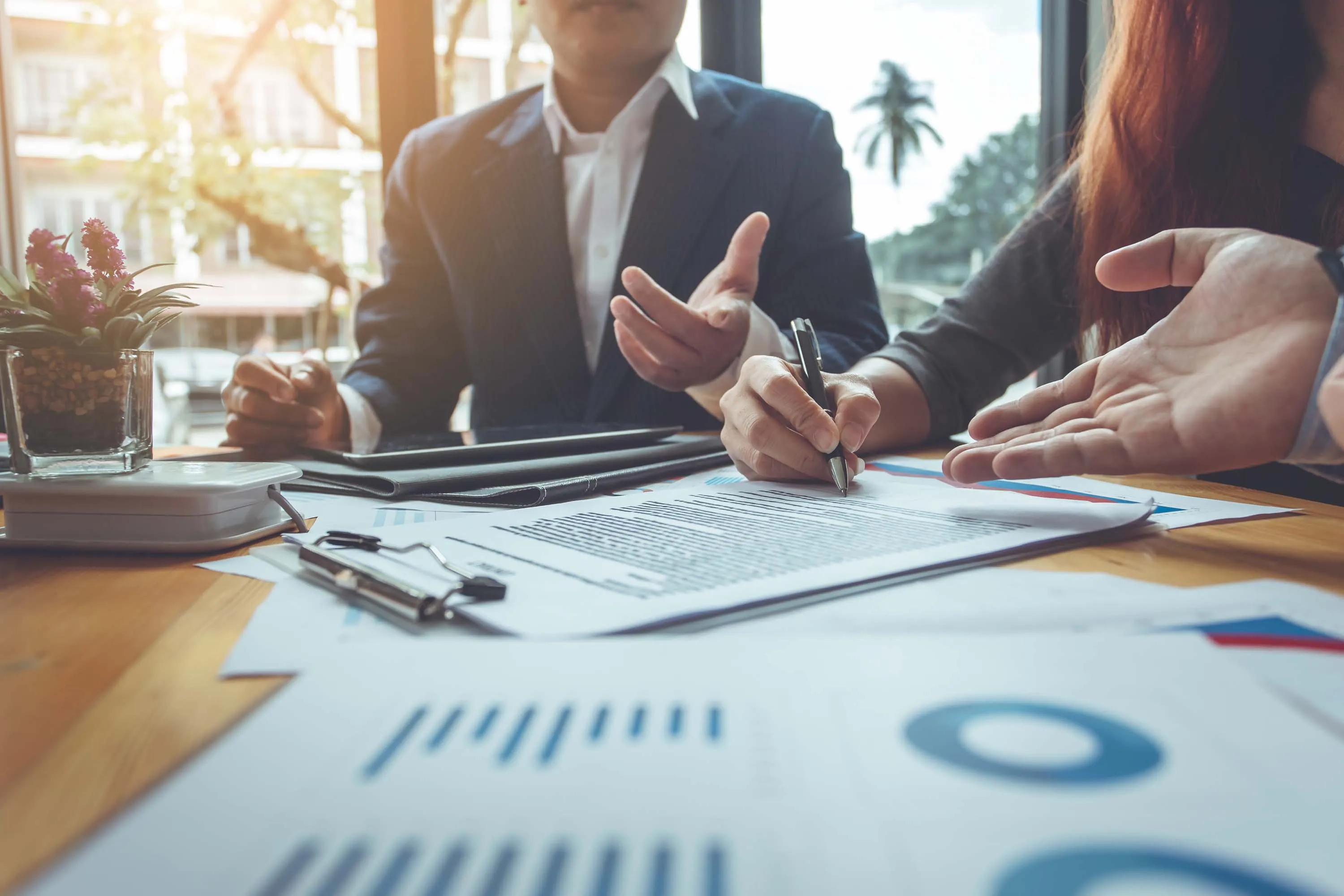 Close-up of a business discussion at a table with documents and charts, one person writing while another gestures with their hand.