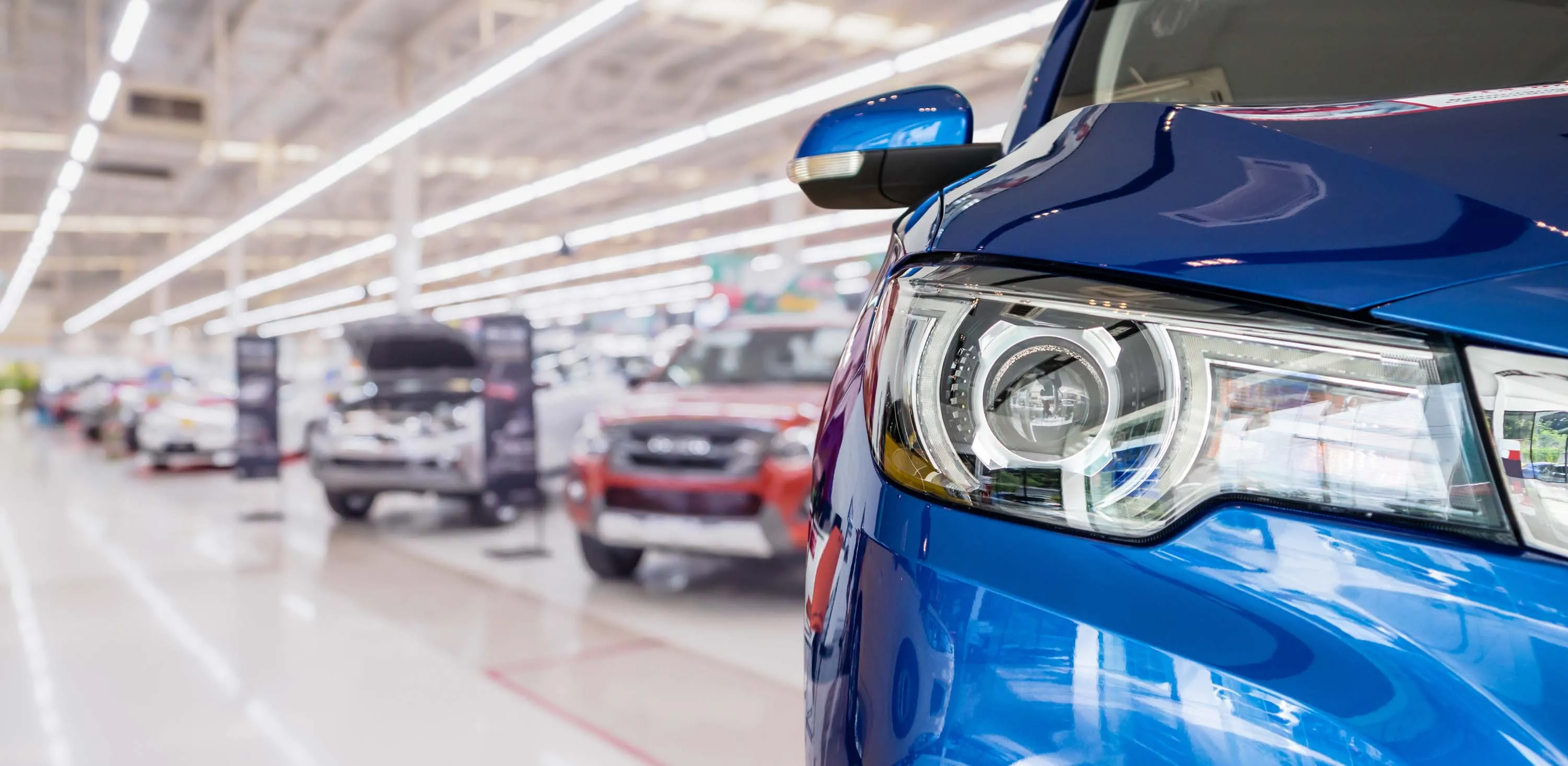 Close-up of a blue car’s headlight inside a brightly lit dealership showroom, with other cars blurred in the background.