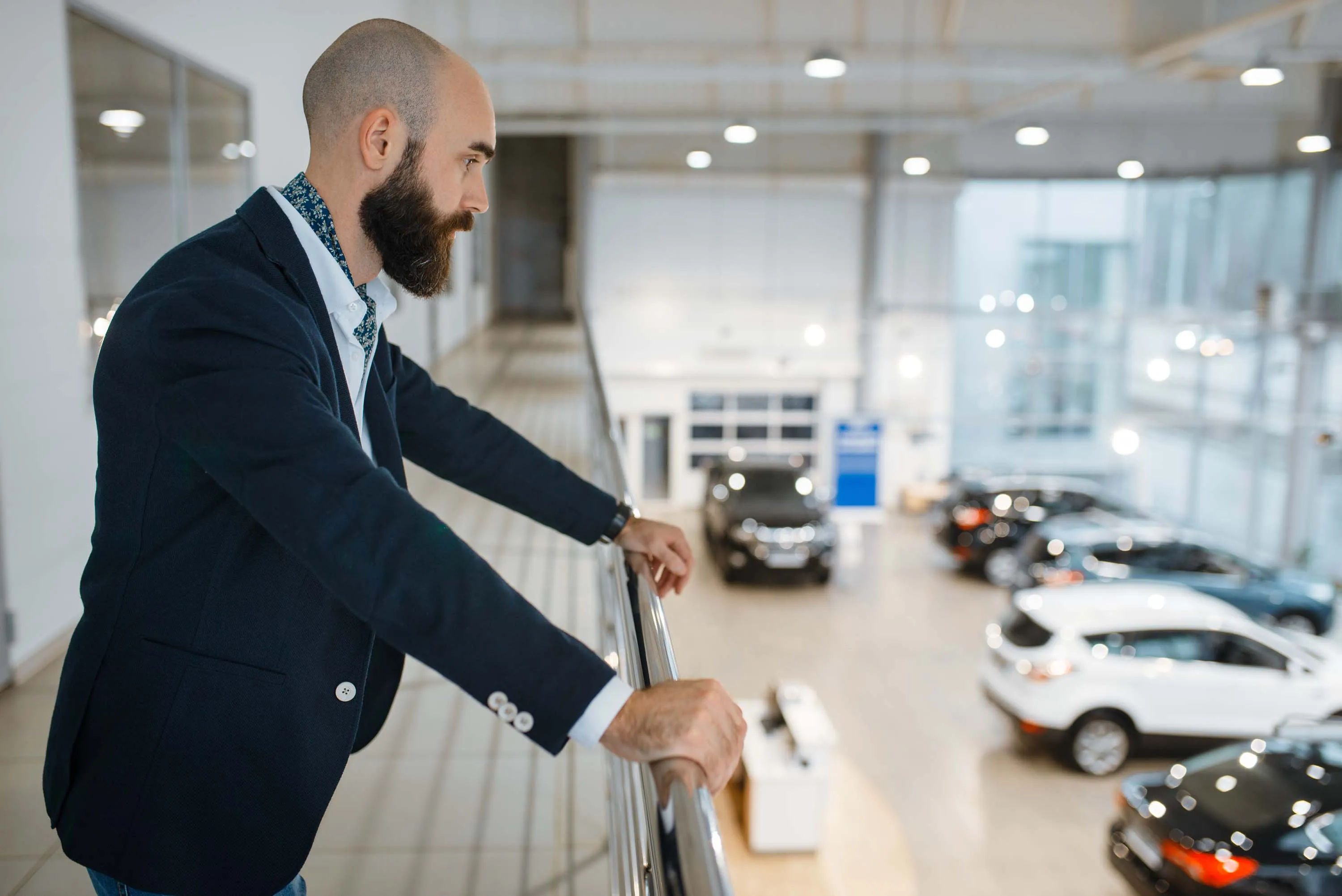 Bearded man in a blazer standing on an upper-level balcony, overlooking cars displayed in a dealership showroom below.