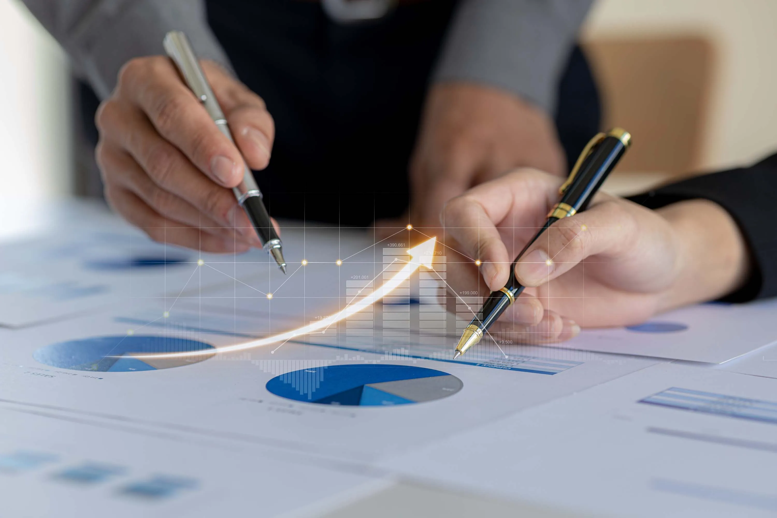 Close-up of two people reviewing financial documents with charts, pointing at graphs with pens, overlaid with a digital graphic of an upward arrow and bar chart.