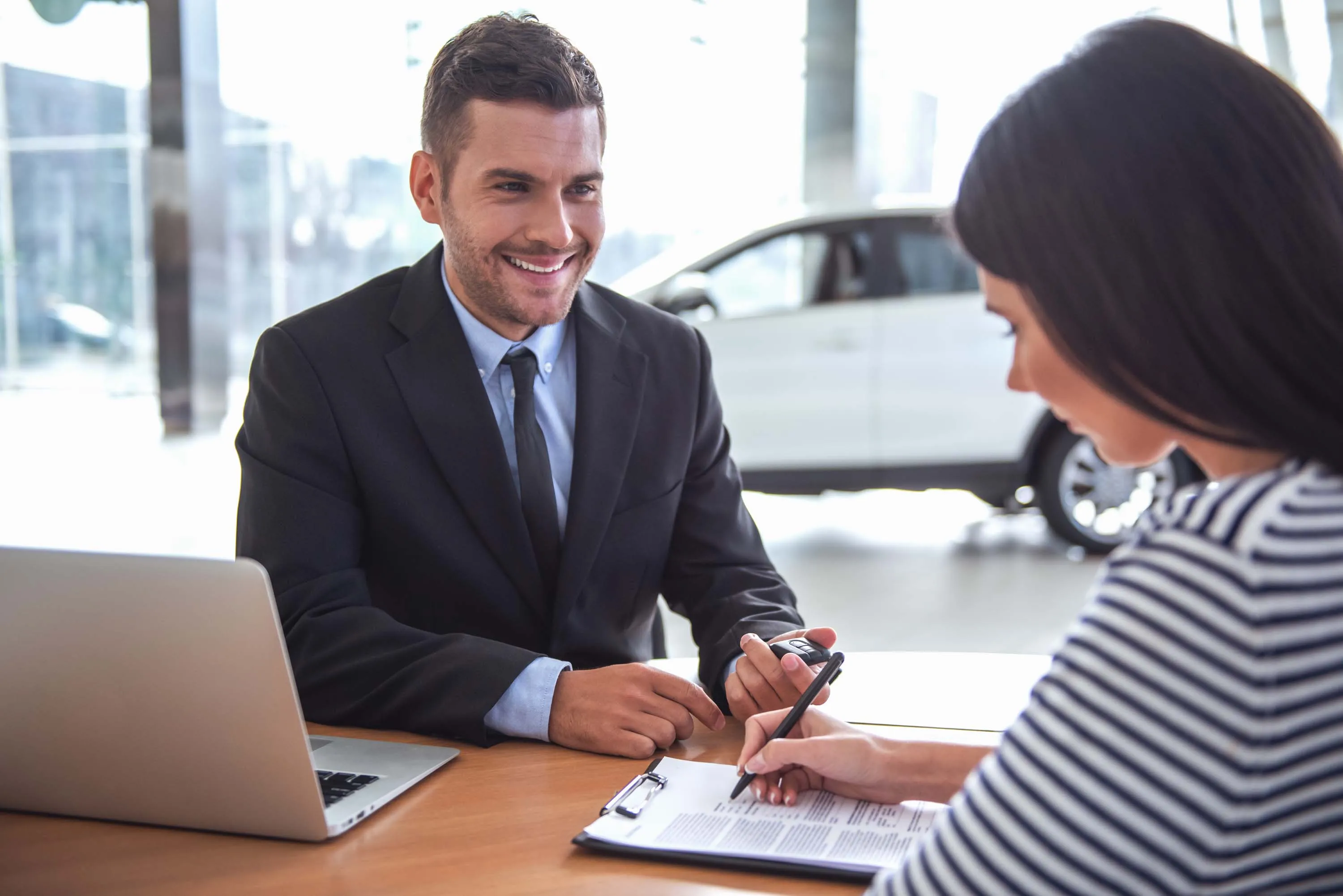Car sales representative in a suit smiling as a customer signs paperwork at a desk, with a laptop open and a car visible in the background.