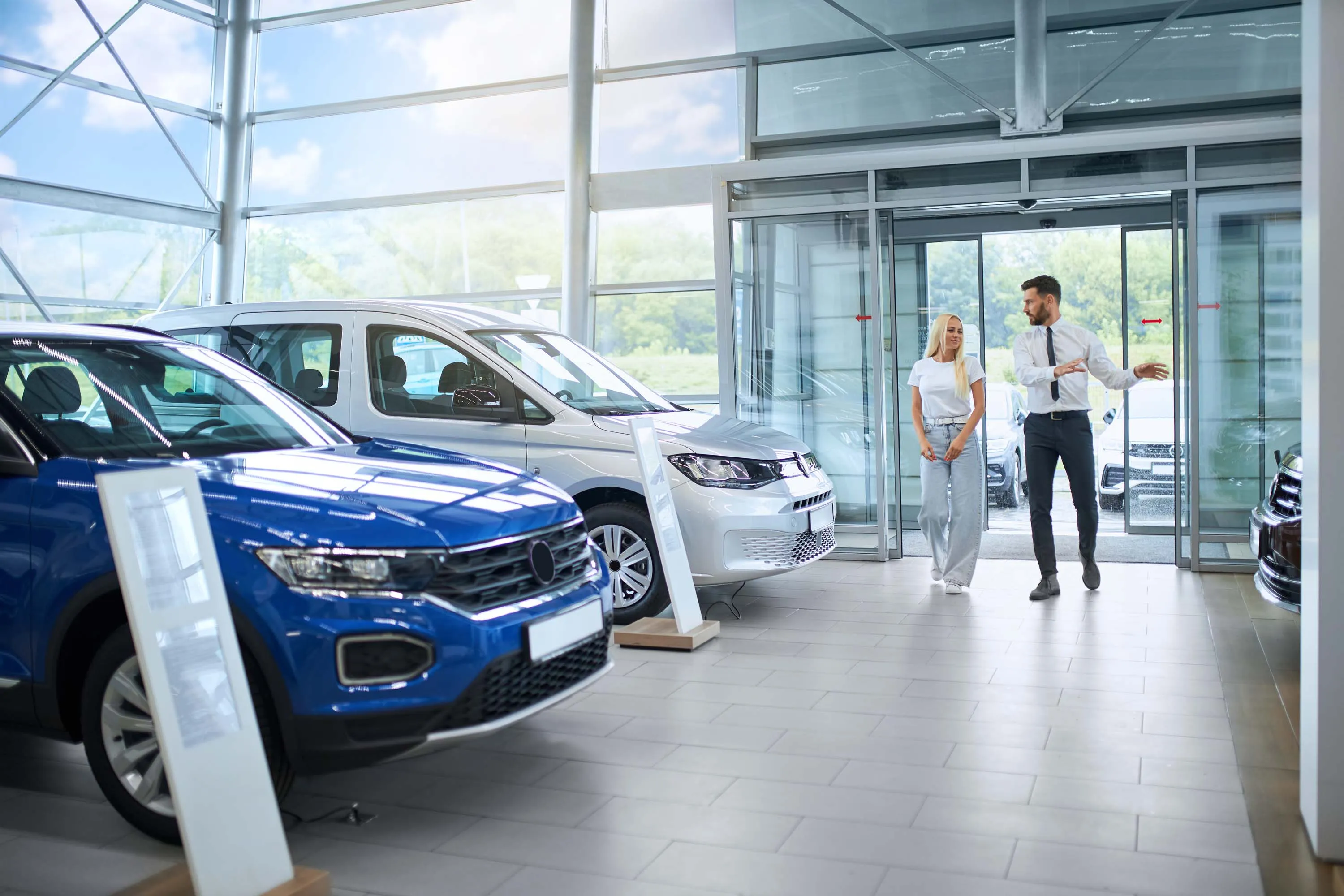 Car salesperson showing a female customer around a dealership showroom with several cars on display, including a blue SUV and a silver van.