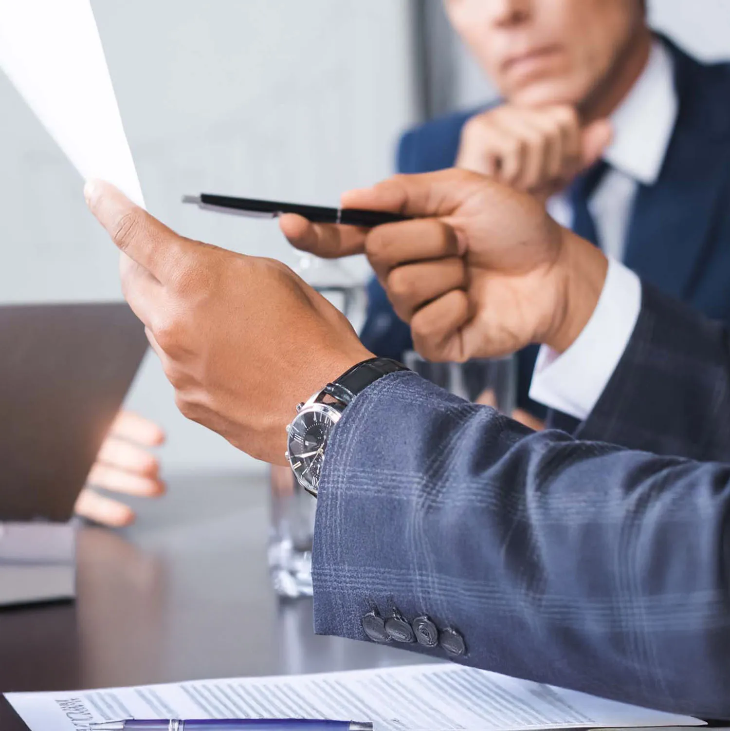 Close-up of a business meeting where a person in a suit is holding and pointing at a document, with water bottles and laptops on the table.