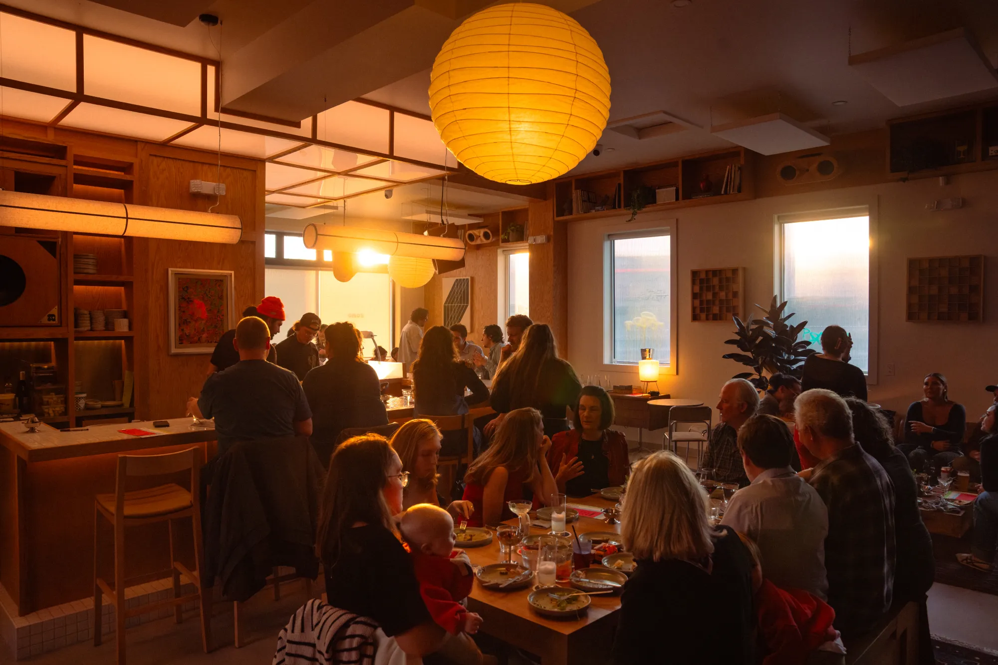 Warmly lit lounge with a large group of people dining and socializing around tables at sunset.