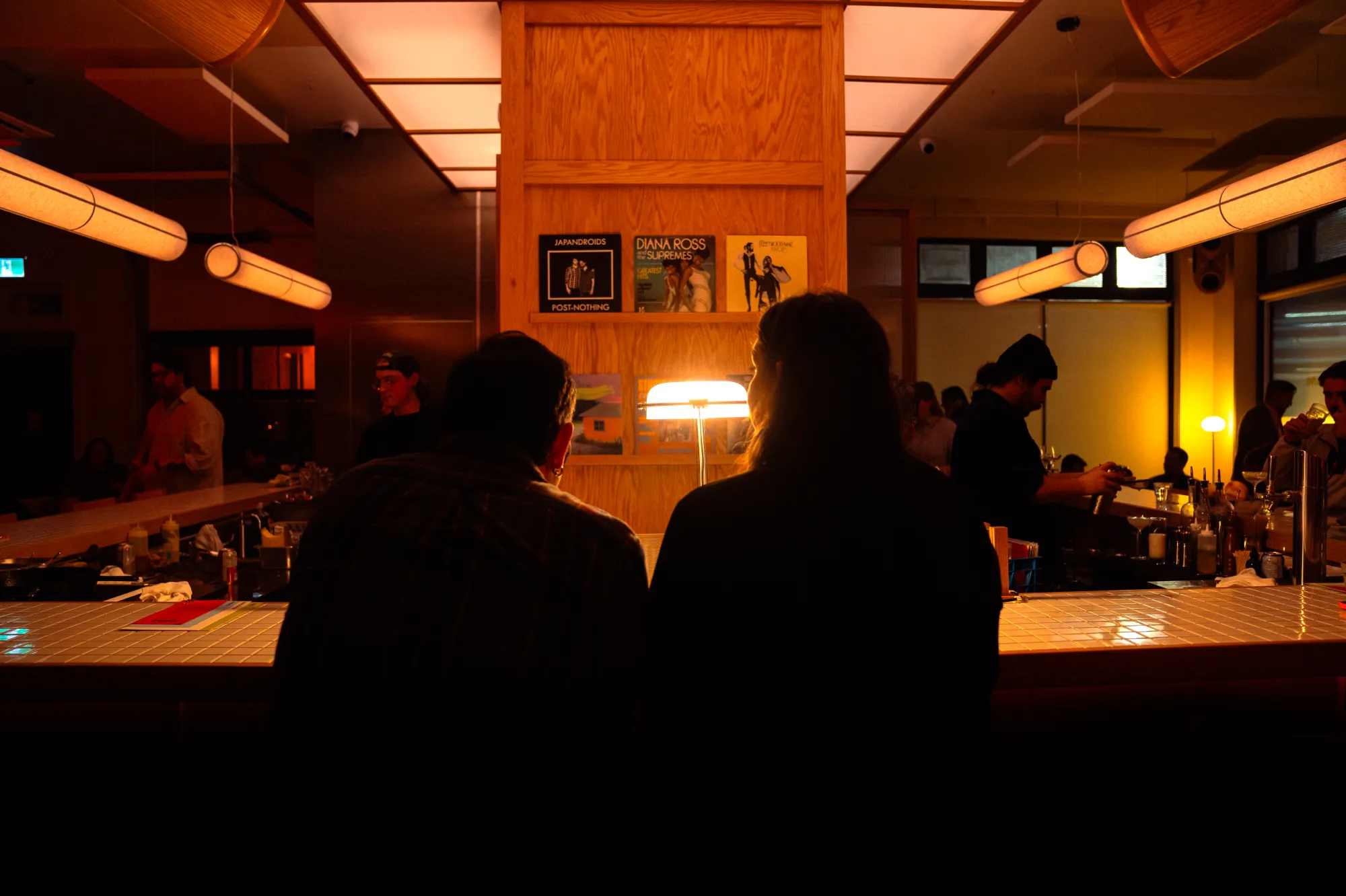 Two people sitting at a bar counter illuminated by warm light in a cozy lounge.