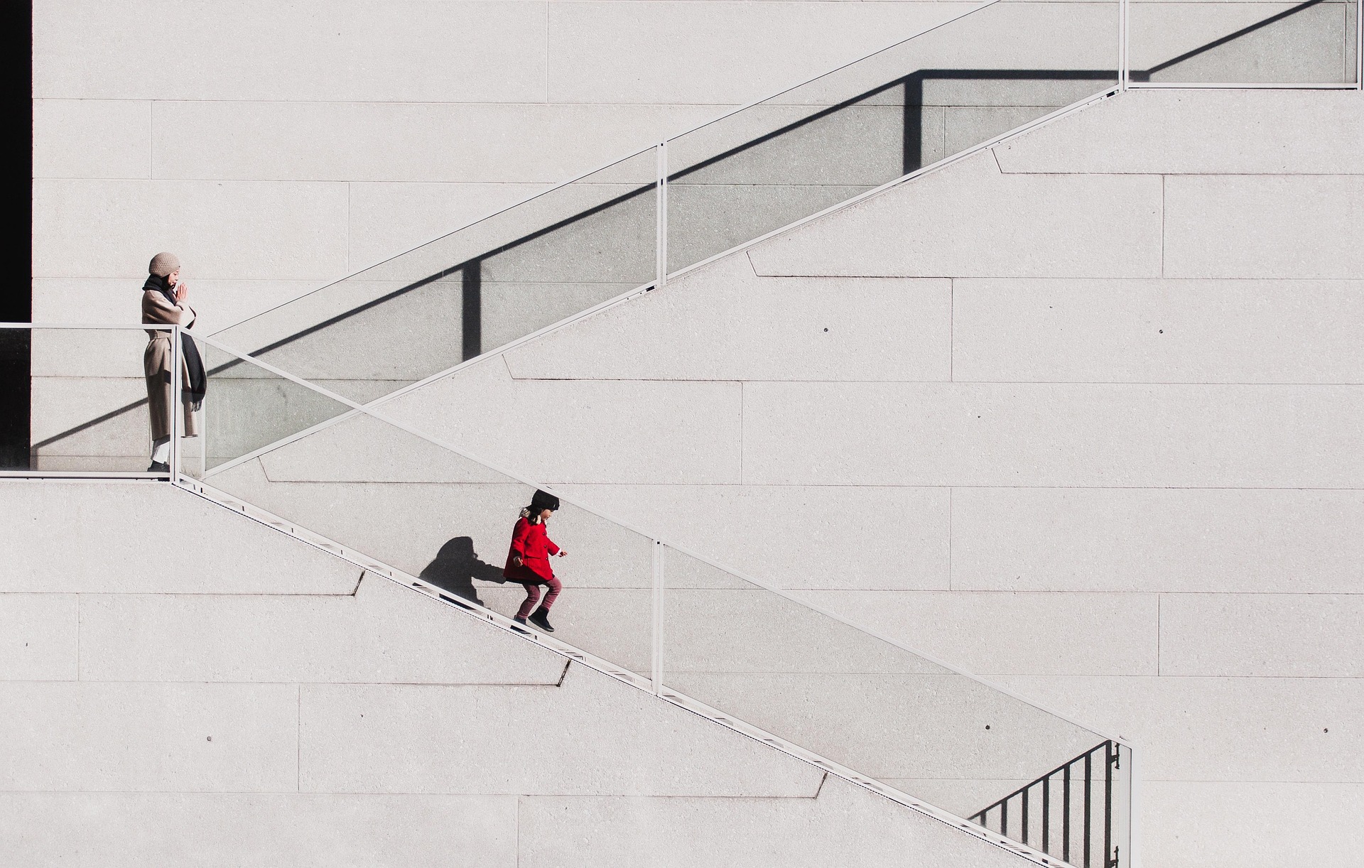 Child in a red coat running down an outdoor staircase while an adult watches from the top landing against a minimalist light gray wall.