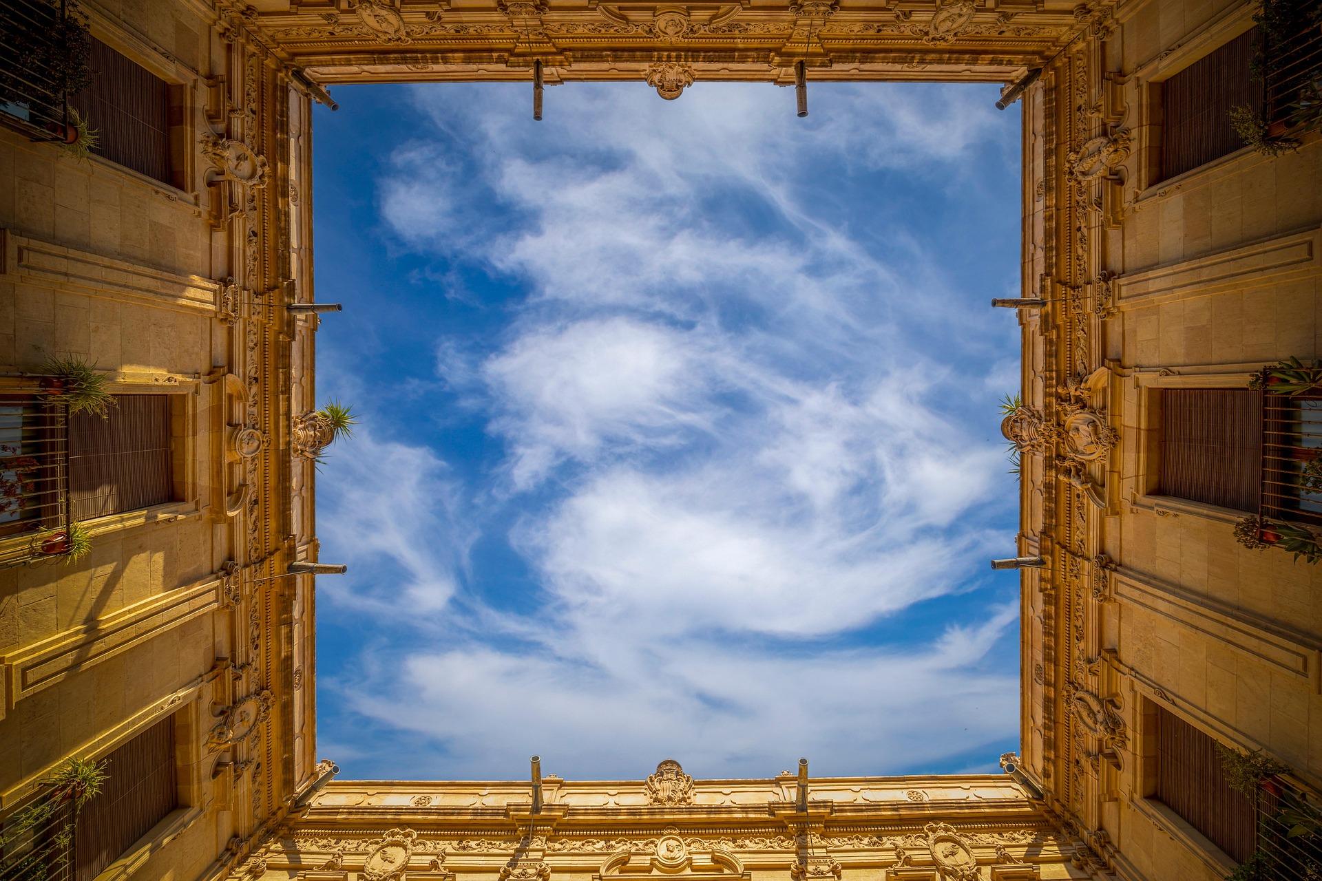 View looking up at a bright blue sky with wispy clouds framed by ornate golden-colored building walls.