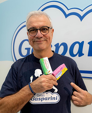 Smiling man with gray hair and glasses wearing a Gelati Gasparini t-shirt holding and pointing at three colorful ice cream bars.