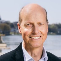 Smiling middle-aged man with a bald head wearing a dark blazer and white shirt outdoors near water.