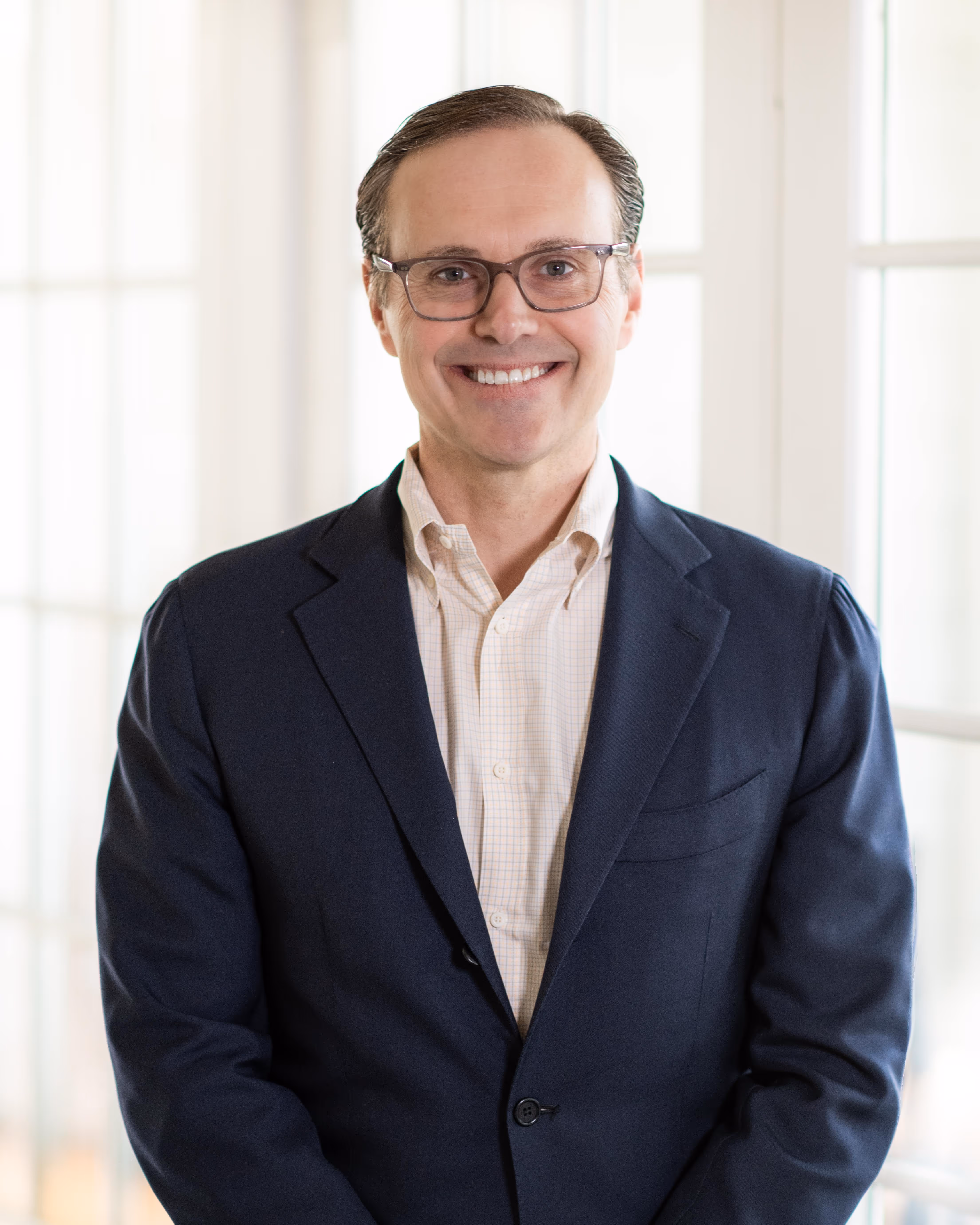 A man in a suit and glasses standing in front of a window.