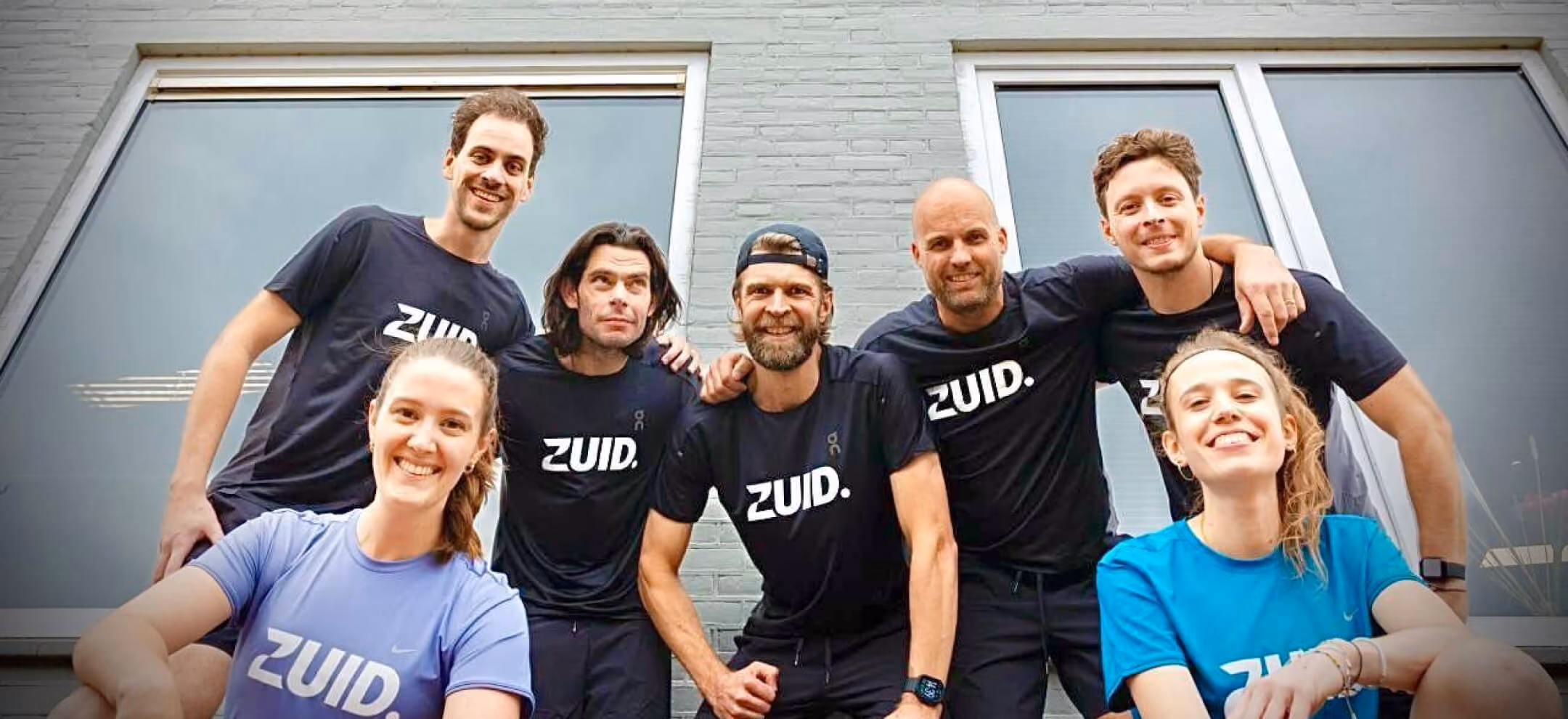 A group photo of seven smiling members of the ZUID. agency team, wearing matching athletic t-shirts with the "ZUID." logo, posed together outdoors in front of a building window.