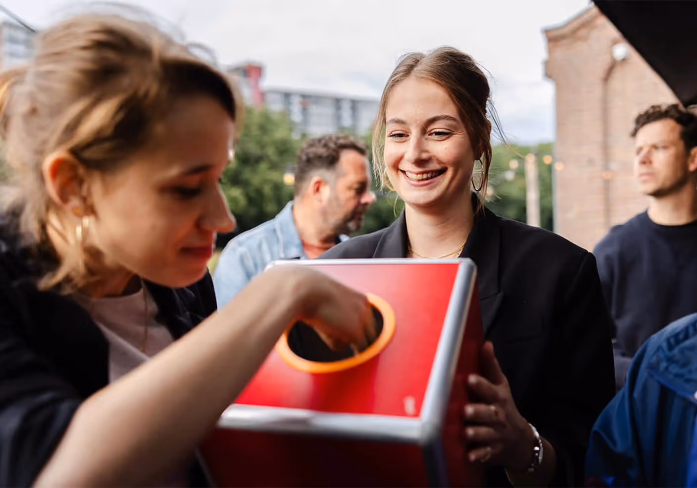Two women from the SOUTH. agency laughing during an outdoor team event. One woman holds a bright red prize draw box while the other reaches her hand inside the circular opening to pull out a ticket or prize.
