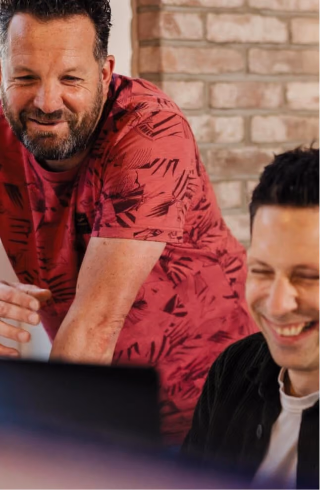 A close-up shot of two male colleagues from the ZUID. agency smiling and laughing together while looking down at a computer screen. One man stands leaning forward in a patterned red shirt, while the other sits in the foreground against a light brick wall backdrop.