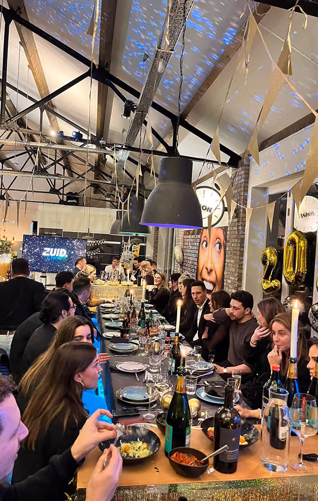 A large group of people from the SOUTH. agency sitting along a long, elegantly set dinner table at a festive event. The scene features warm candle lighting, wine bottles, and food, under an industrial ceiling decorated with gold pennant banners. A digital screen displaying the ZUID. logo is visible in the background.