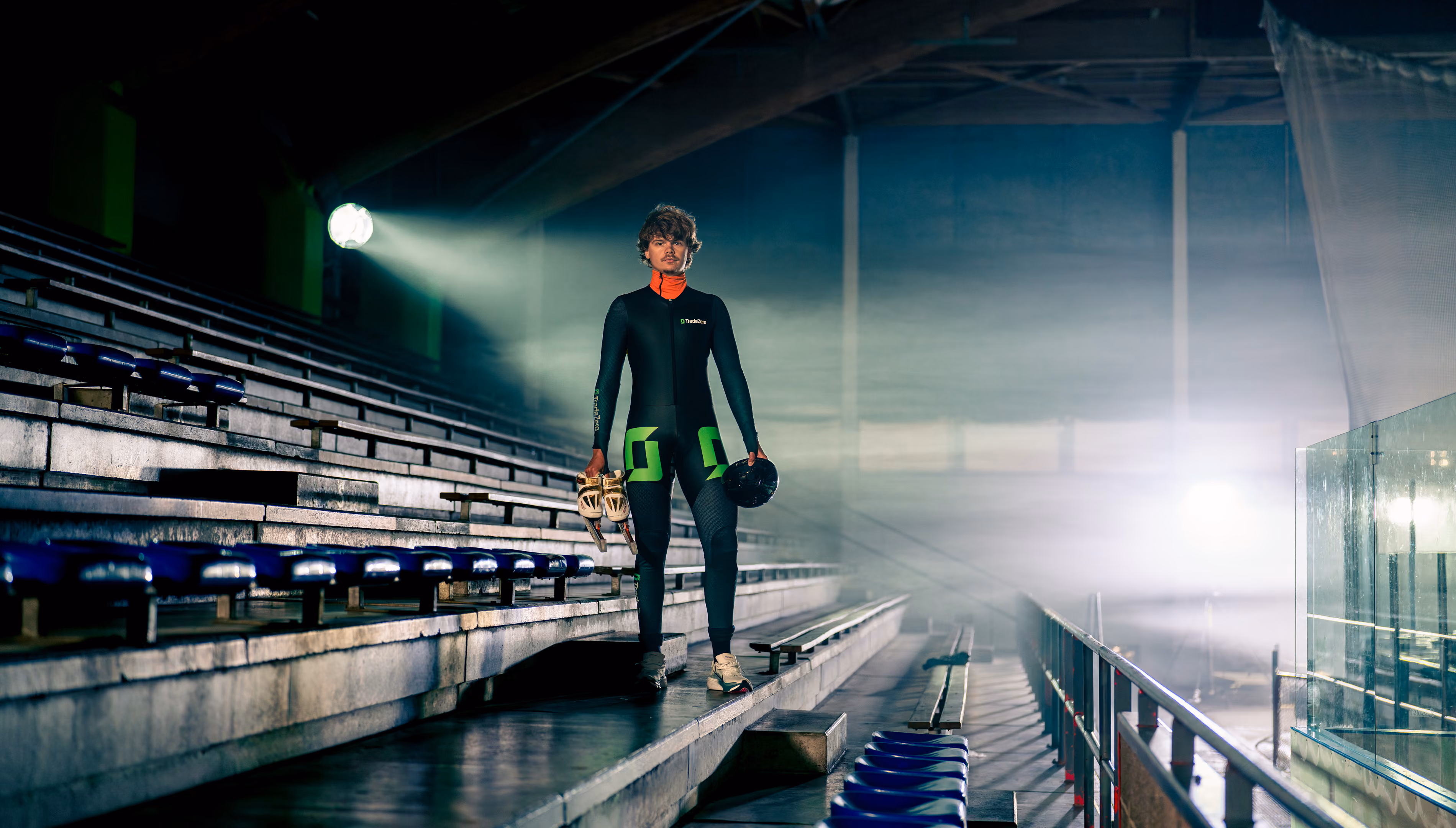 IJschaatser in zwart pak met groene accenten staat in lege tribune en houdt schaatsen en helm vast.