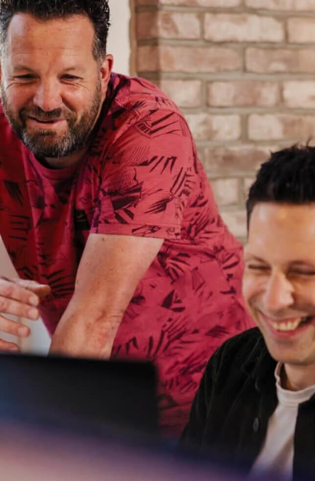 Twee mannen lachen en kijken samen naar een laptopscherm in een informele werkomgeving.