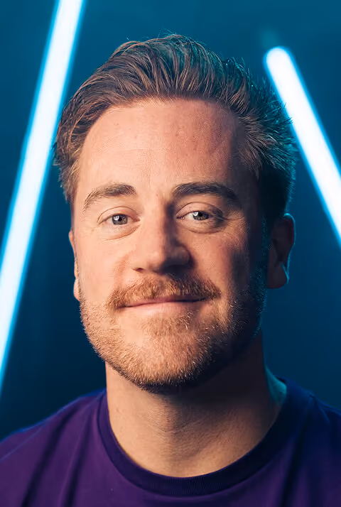 Portret van een glimlachende man met lichtbruine baard en blauwe ogen, met vage blauwe neonlichten op de achtergrond.
