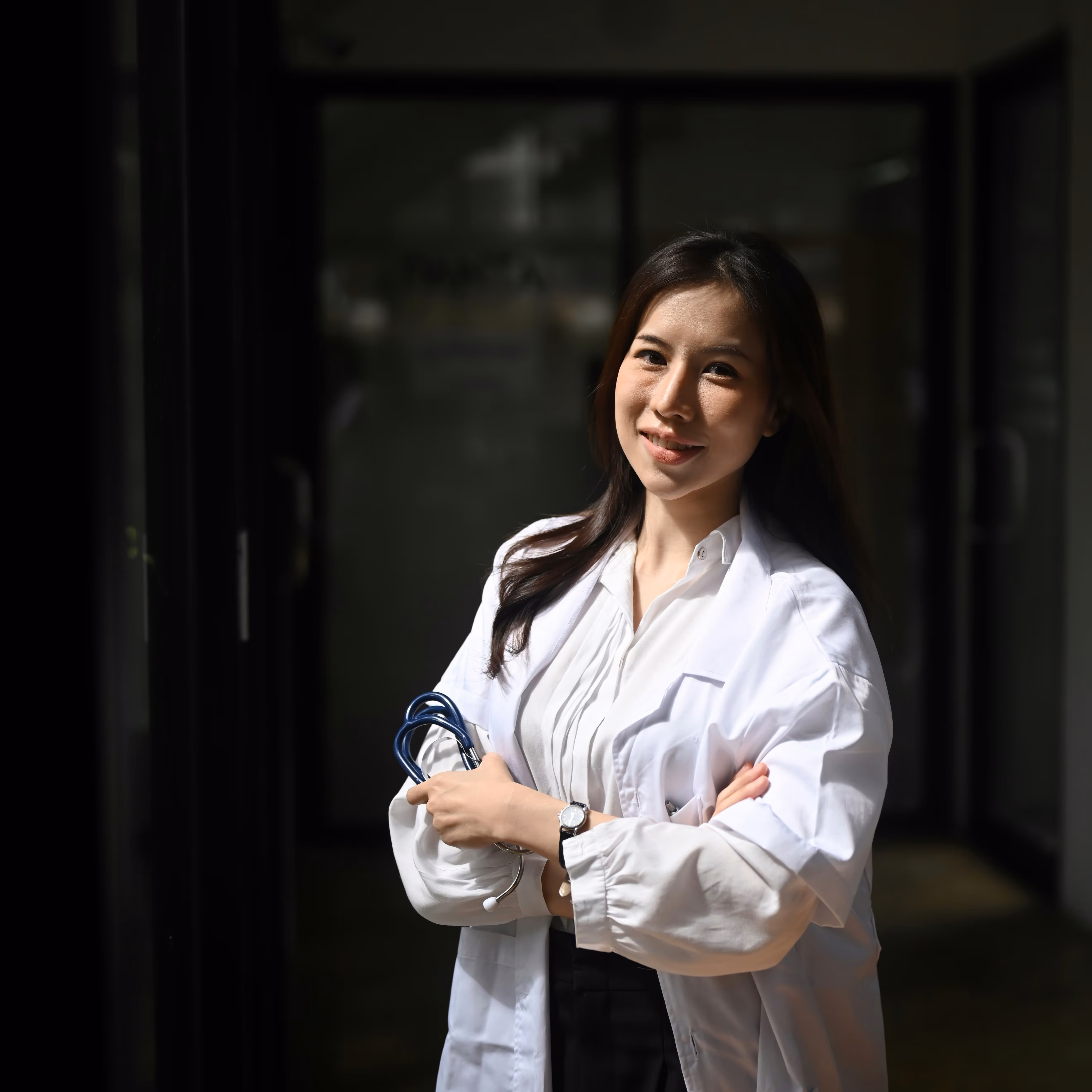 Vrouwelijke arts glimlachend met gestifte armen en stethoscoop in witte jas in een donkere gang.