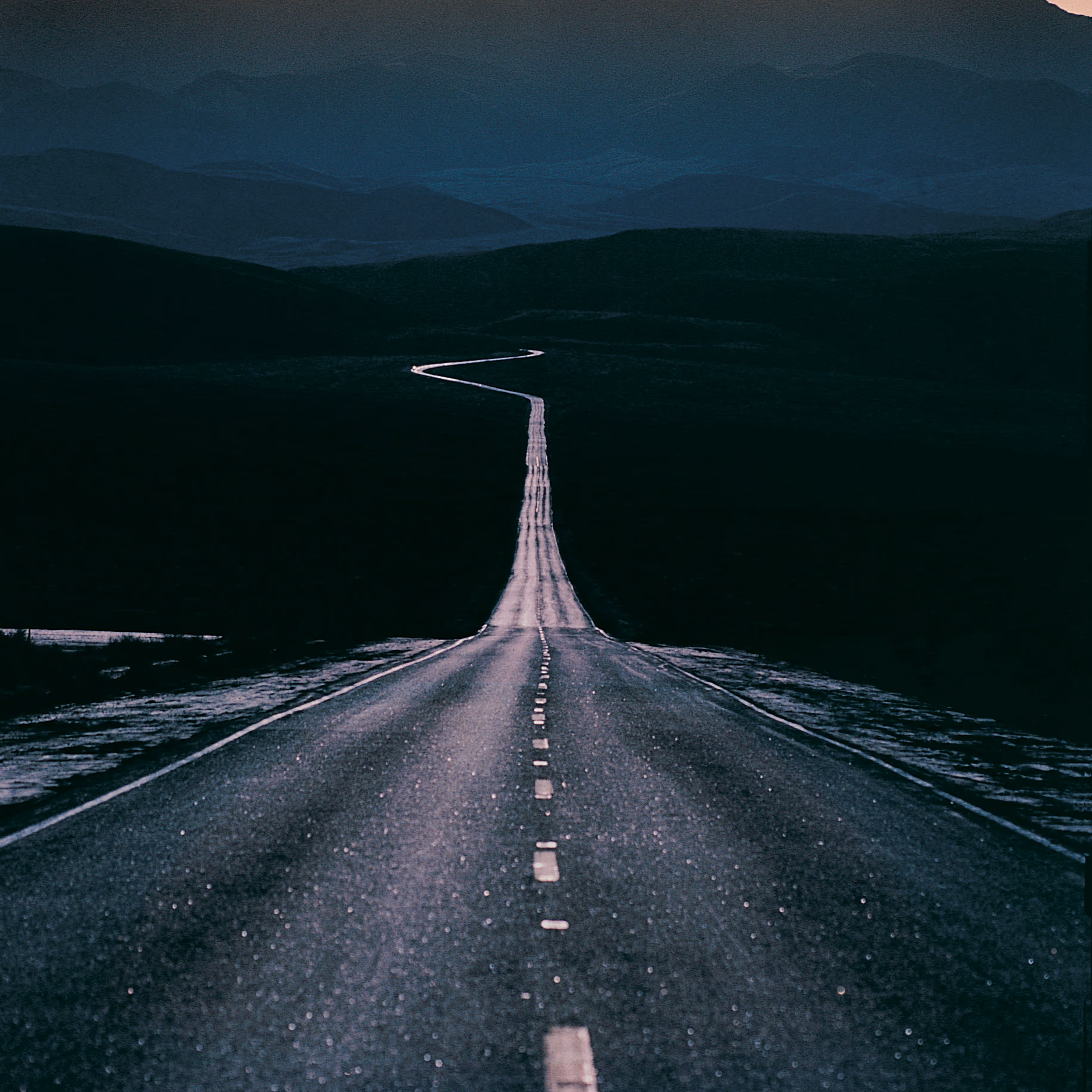 Lang, recht stuk weg dat overgaat in een kronkelende weg door een donker berglandschap bij schemering.