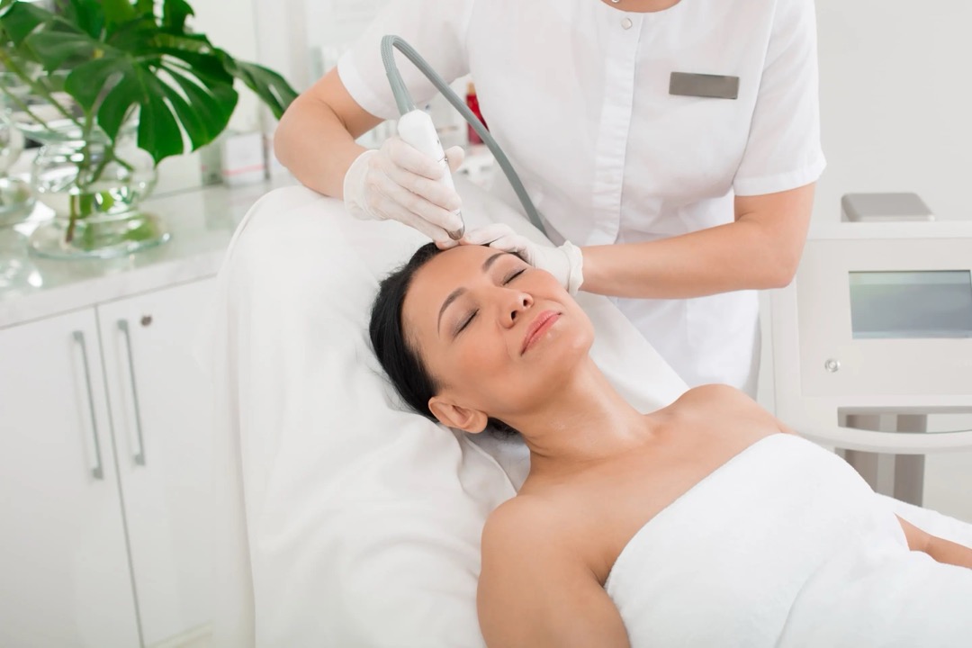 A woman lies back with her eyes closed while a gloved clinician holds a laser handpiece near her forehead