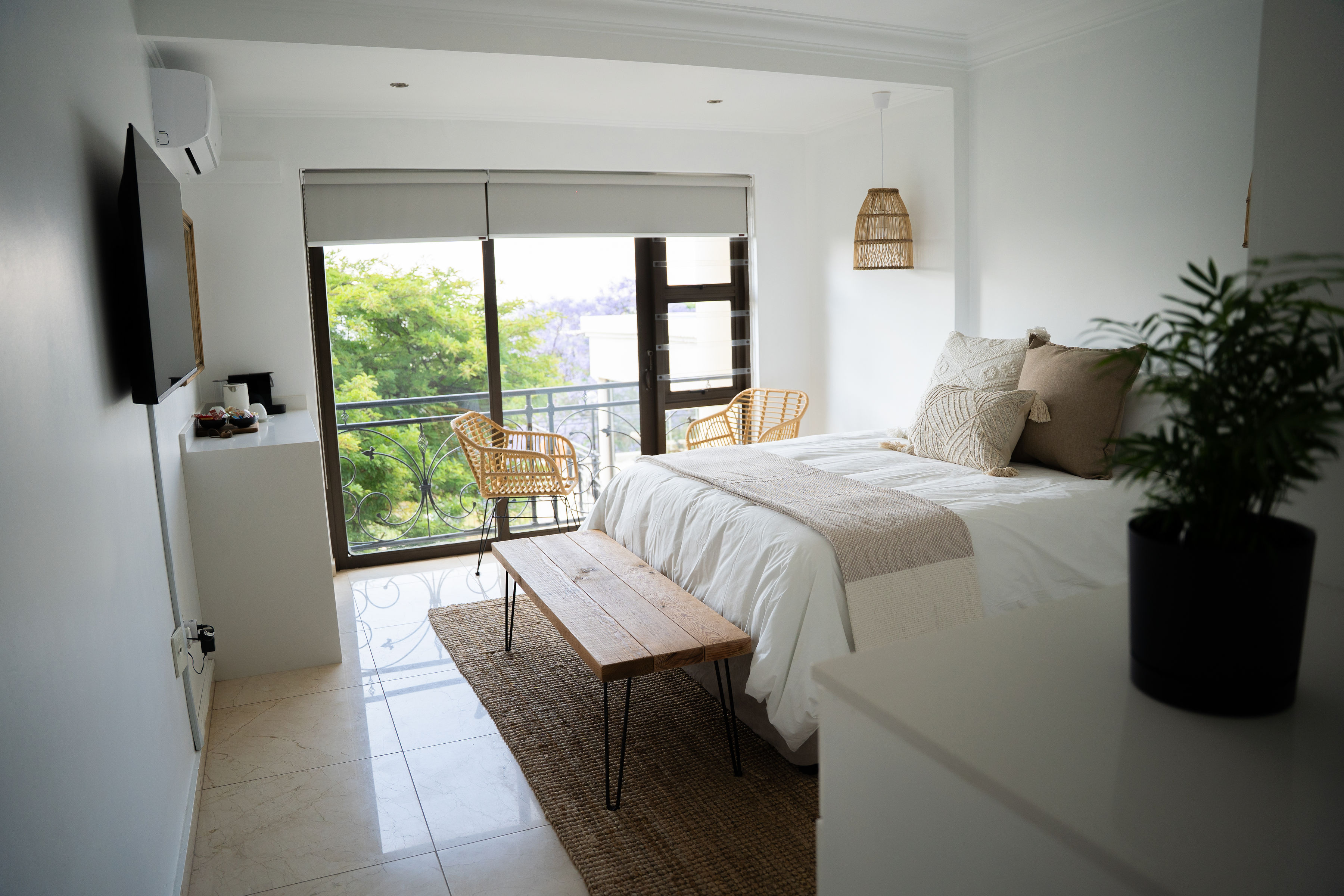 Modern bedroom with white walls, a bed with beige and white pillows and throw, wooden bench, two wicker chairs by large window, and a potted plant.