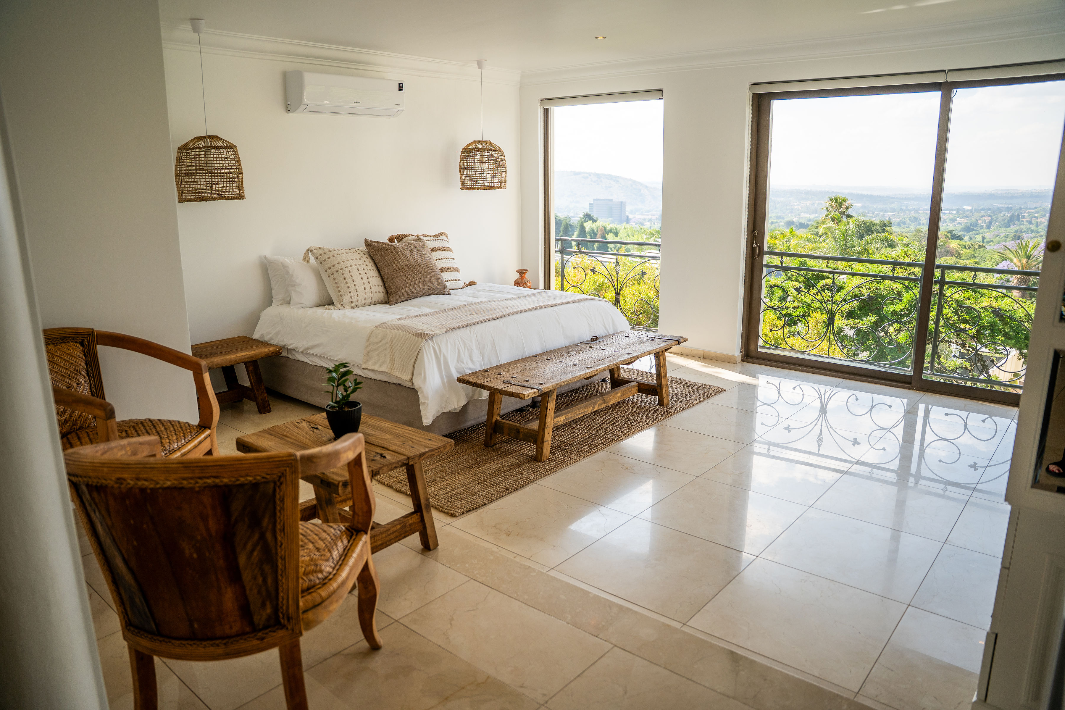Bright bedroom with white walls, a bed with beige and brown pillows, wooden furniture, and large windows overlooking green trees and hills.