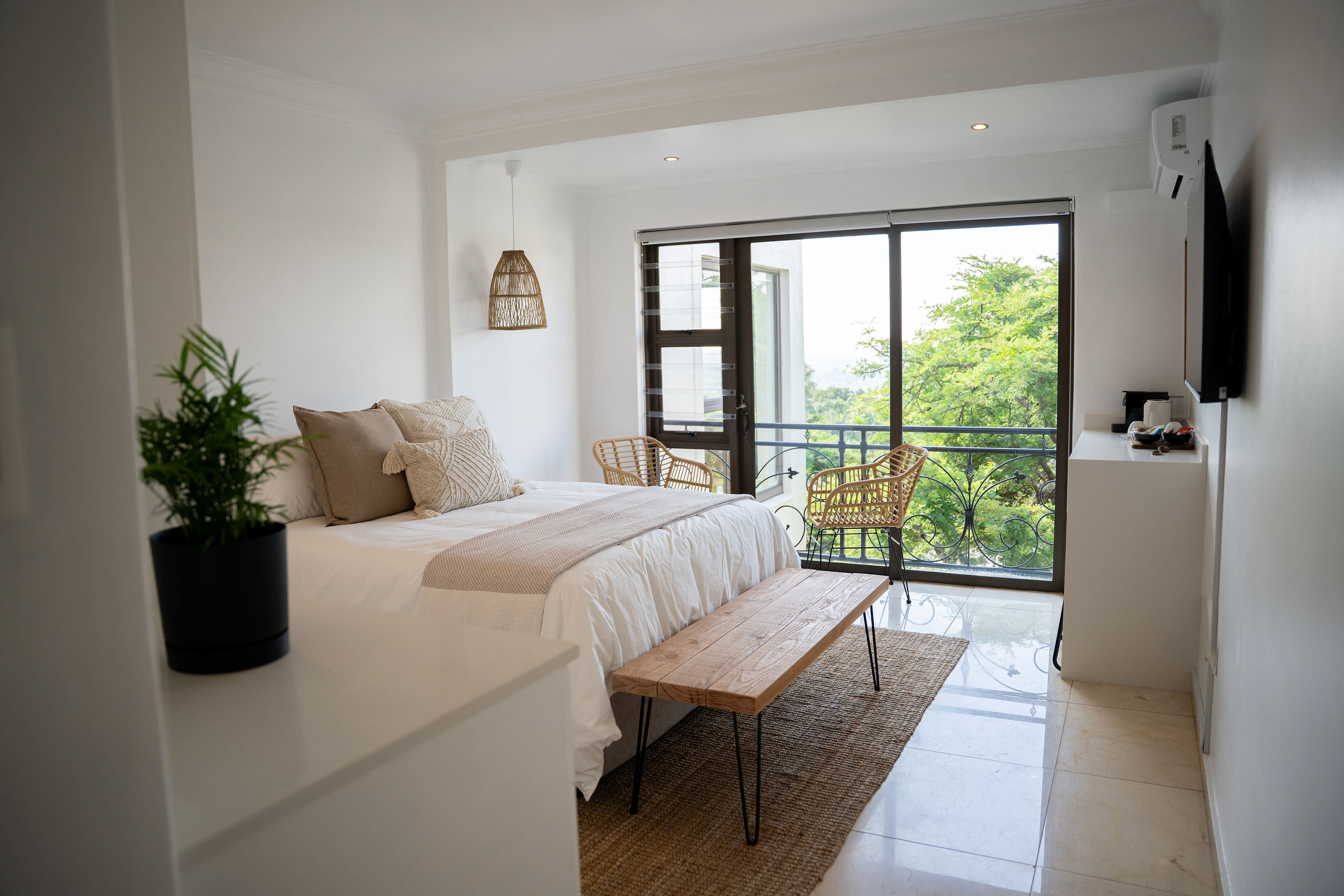 Bright bedroom with a white bed, beige pillows, wooden bench, two wicker chairs near balcony overlooking greenery.