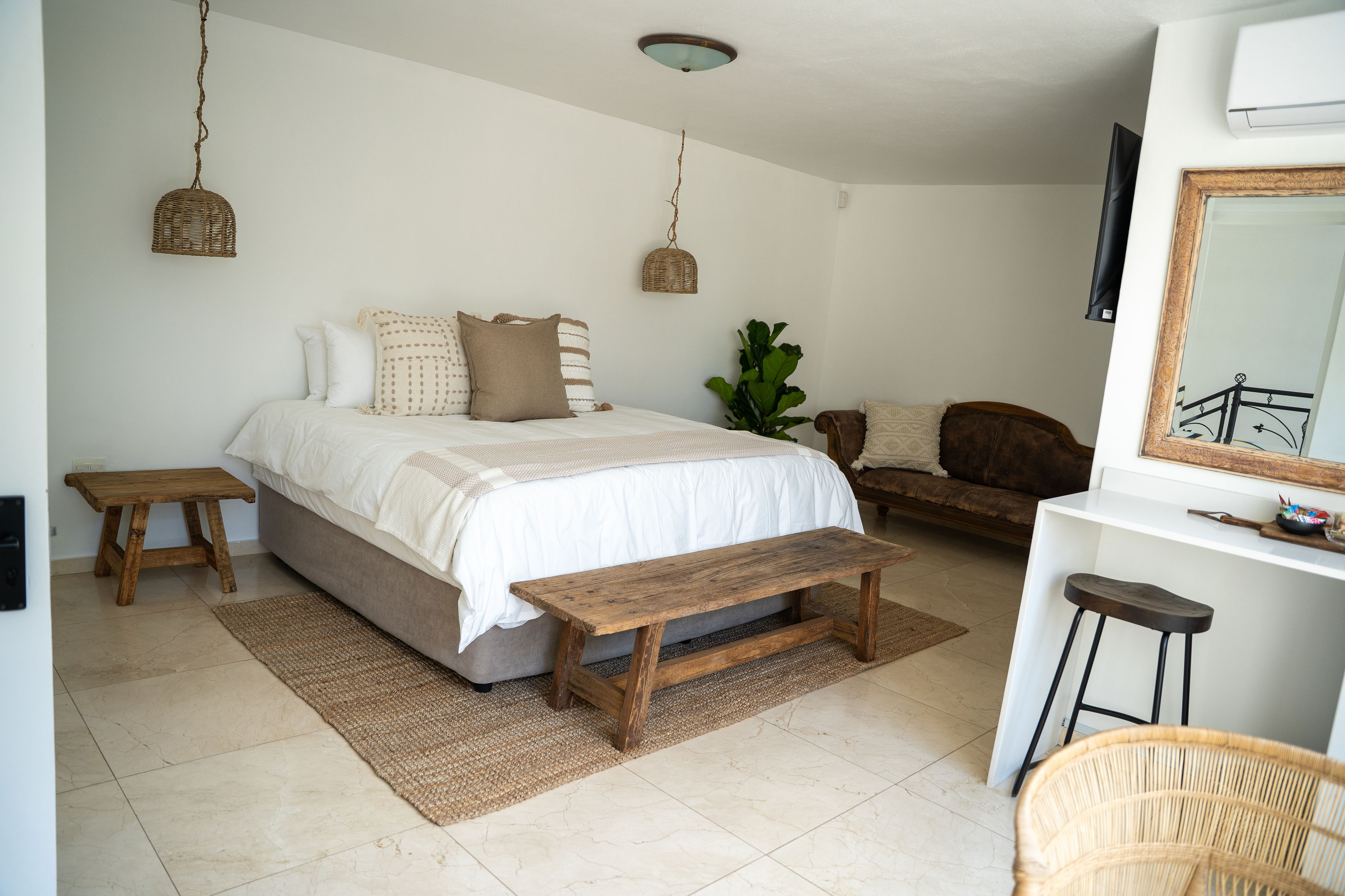 Modern bedroom with a bed featuring white and beige bedding, wooden bench at the foot, wicker pendant lights, and a vintage brown sofa in the corner.