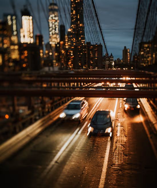 Blurred night view of cars driving on a city bridge with glowing building lights in the background.