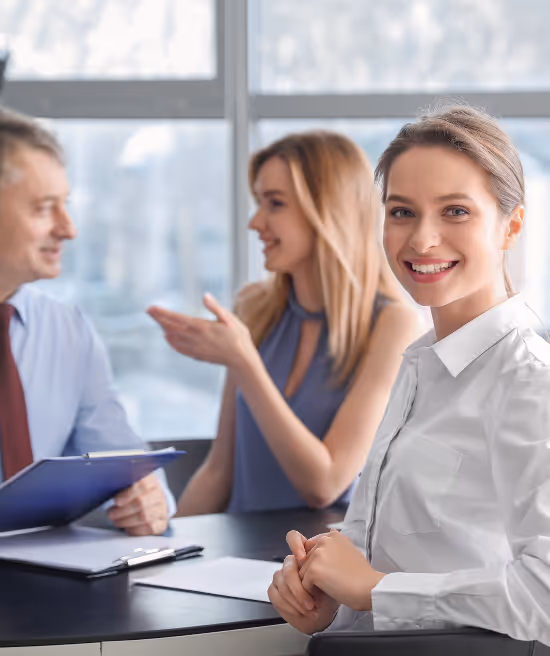 Smiling businesswoman in white shirt sitting at a table with two colleagues engaged in discussion behind her.