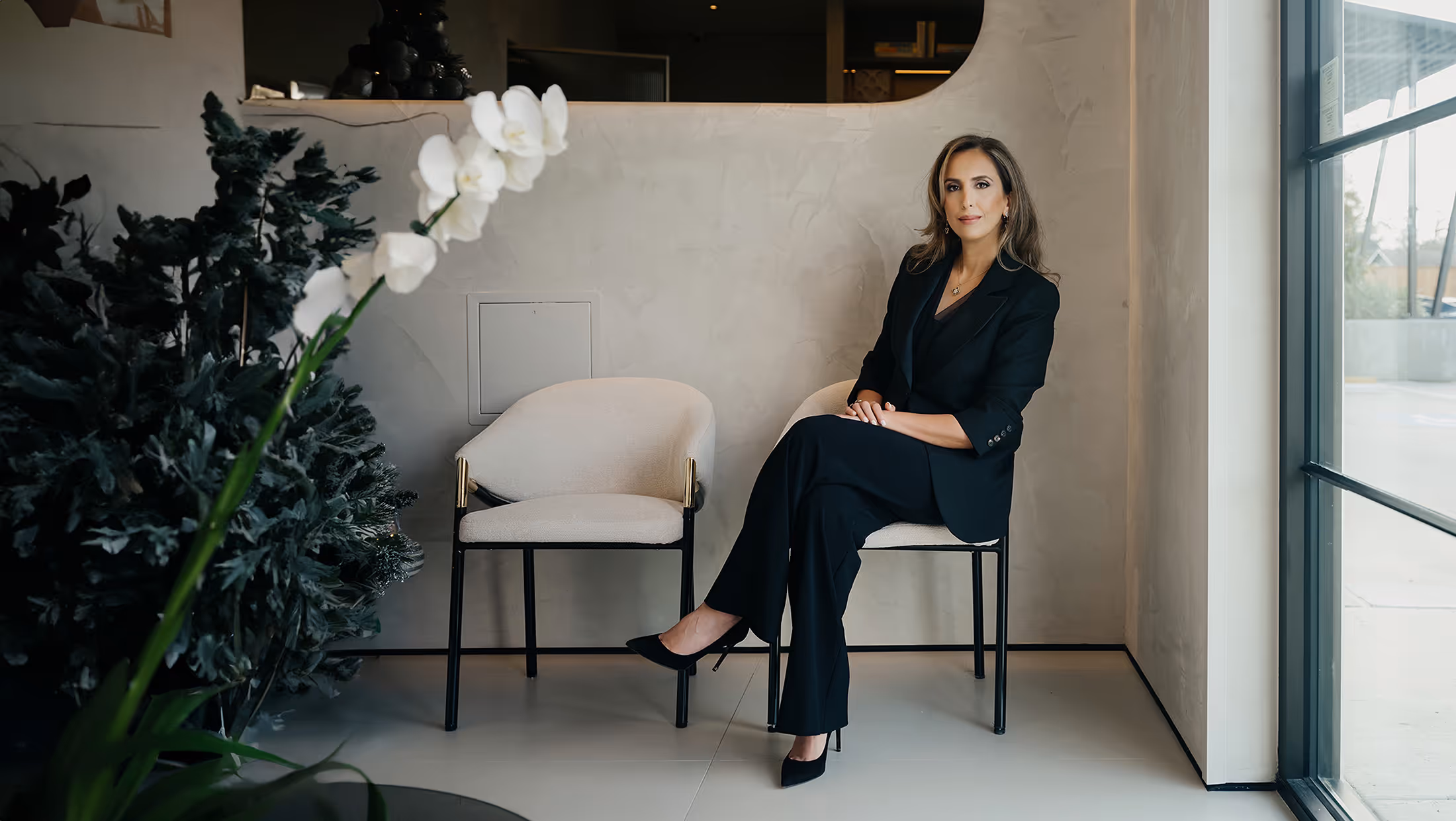 A professional portrait of a woman in a black suit sitting in a modern, minimalist interior setting next to a large potted pl