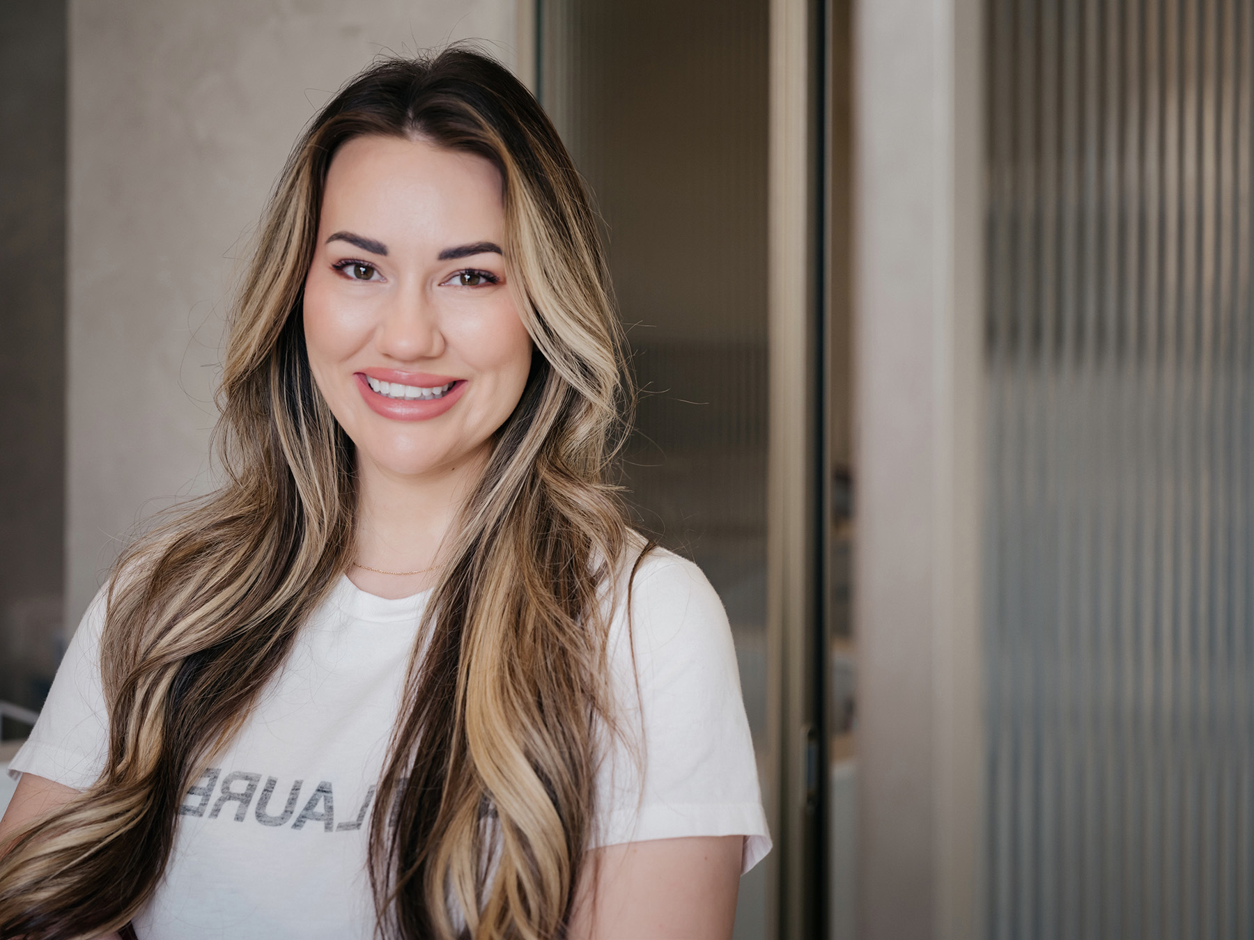 A portrait of a smiling woman with long wavy hair wearing a white t-shirt against a neutral background. — Clio Dental Studio