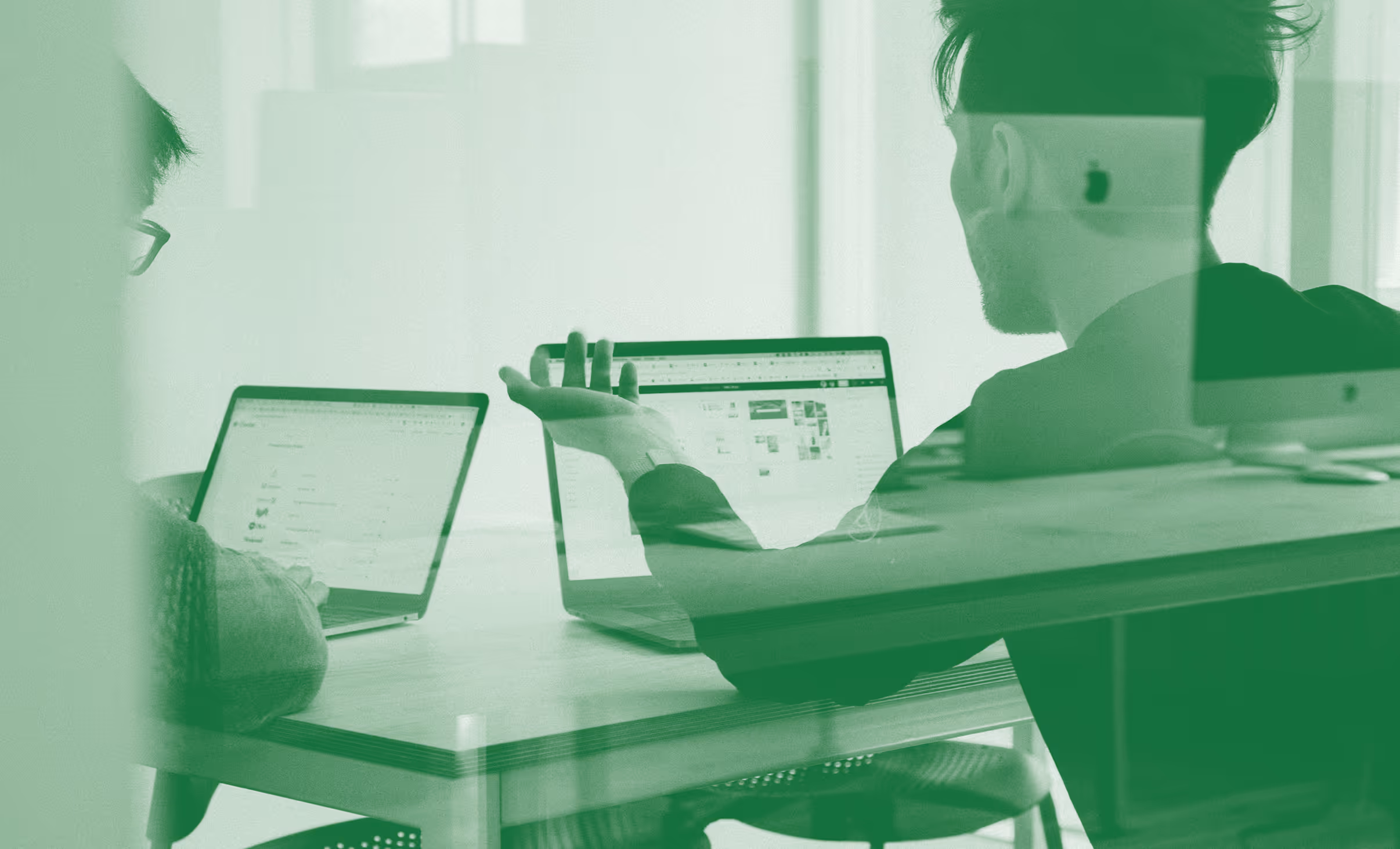 Two people seated at a table working on laptops, one gesturing towards the screen during a discussion.