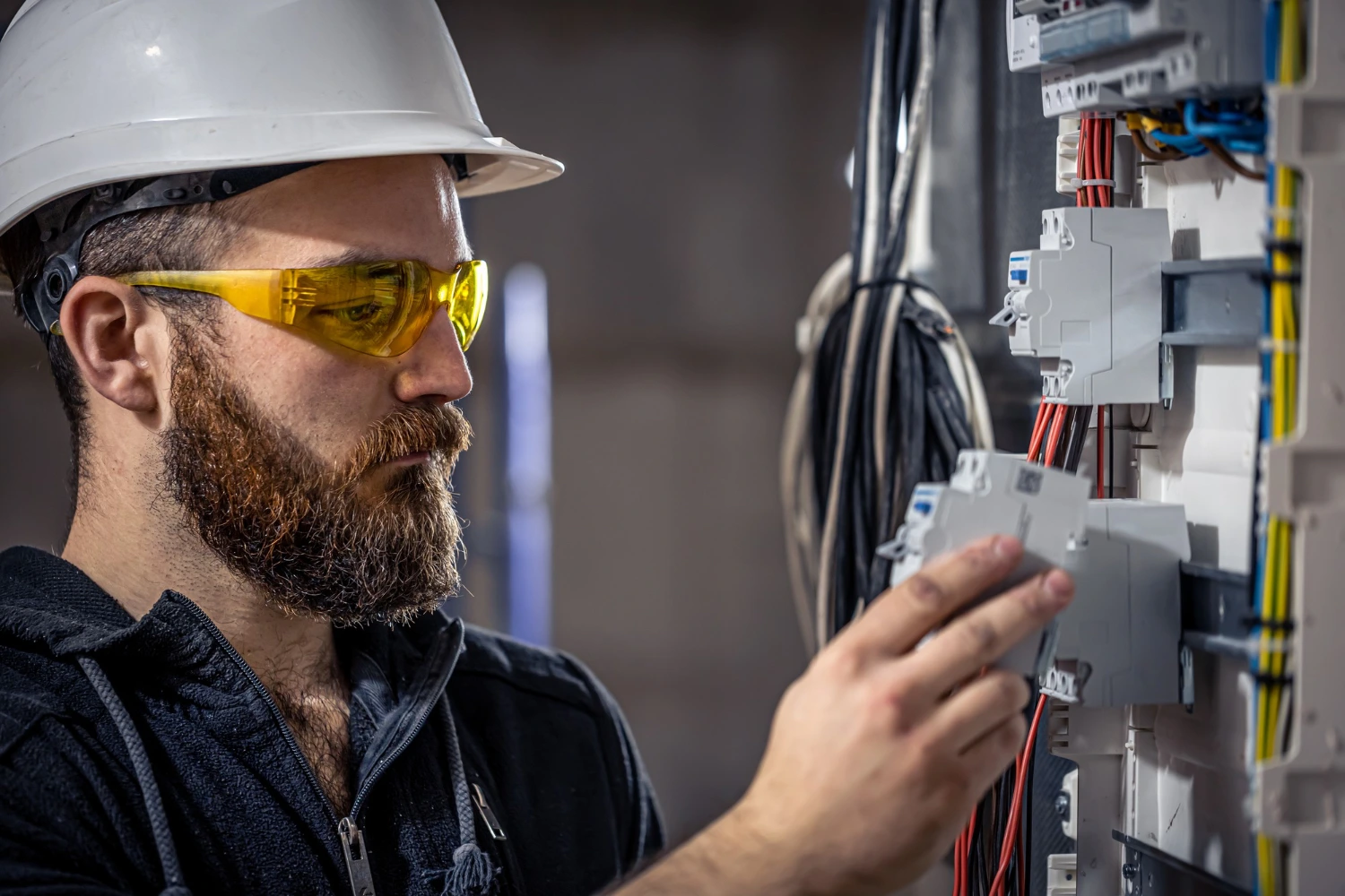 A man wearing a hard hat and safety glasses is inspecting electrical wires on a switchboard.